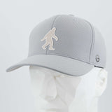 The Eternal Glyph Bigfoot Silhouette Albino Flexfit® Cool & Dry Cap in silver features a bold Bigfoot design on the front and a small folklore-inspired circular logo on the side, displayed on a faceted white mannequin head.