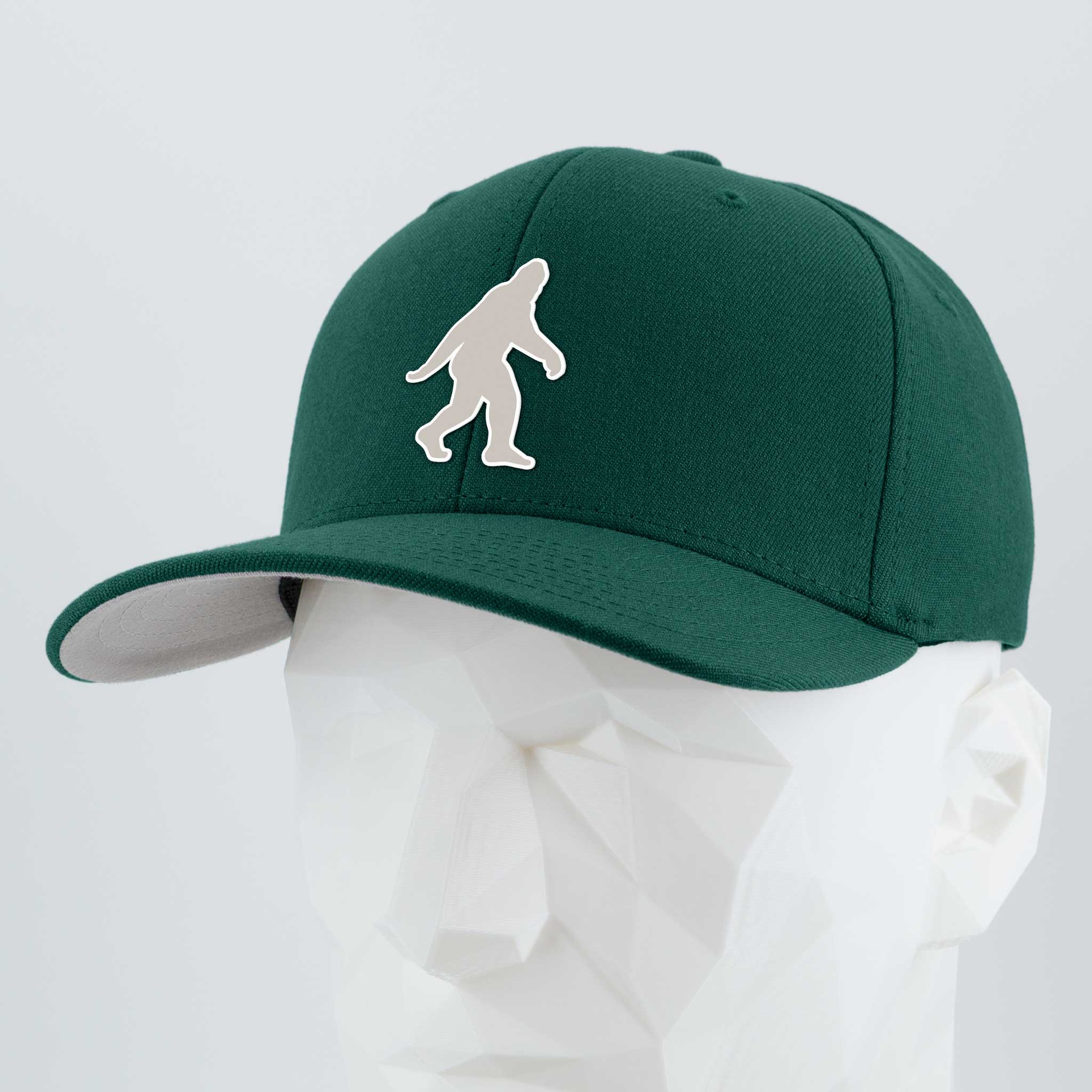 A spruce Eternal Glyph Flexfit® Premium Wool Blend Cap, featuring a white Bigfoot silhouette logo on the front, is displayed on a white geometric mannequin head against a plain light background.