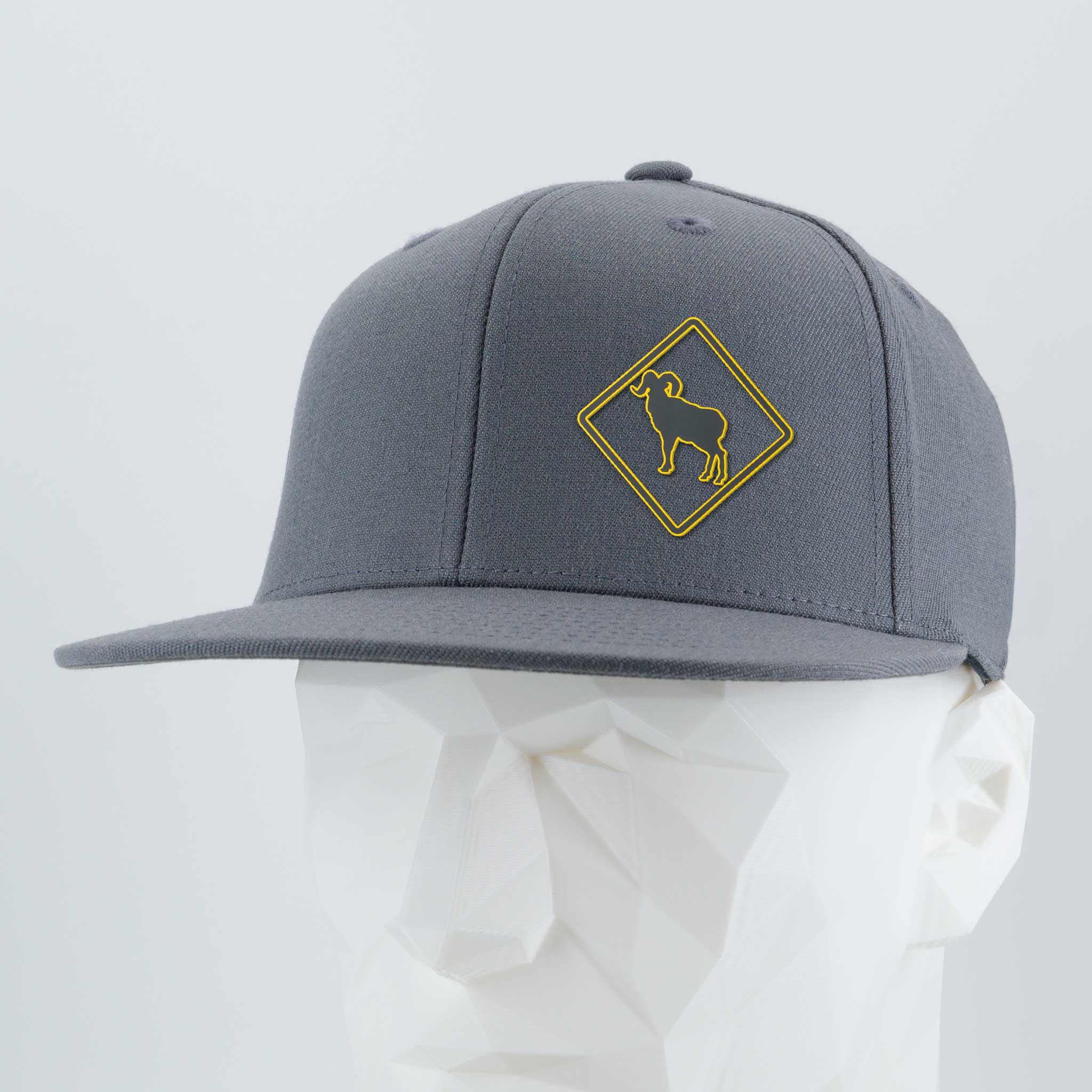 The Eternal Glyph Bighorn Sheep Crossing Sign (Silicone) Flexfit® Flat Bill Premium Fitted Cap is a gray, flat-brimmed hat with a yellow bighorn sheep road sign, shown on a white geometric mannequin head.