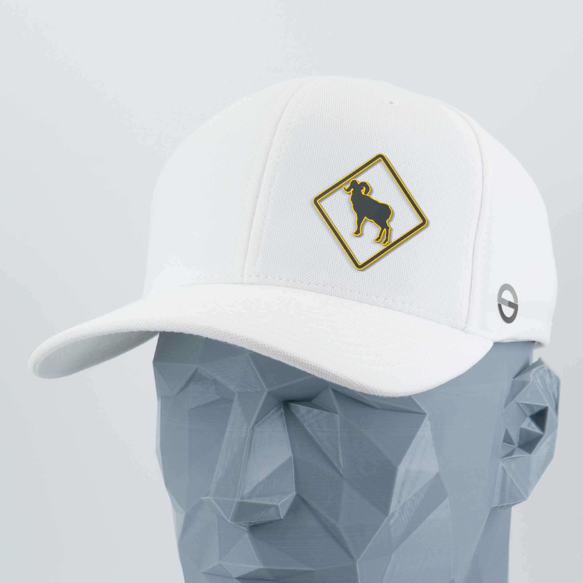 A geometric gray mannequin head displays the Eternal Glyph Bighorn Sheep Crossing Sign (Silicone) Flexfit® Cool & Dry Cap—white, with a yellow-bordered patch showing a black bighorn sheep silhouette and a small gray circular side logo.