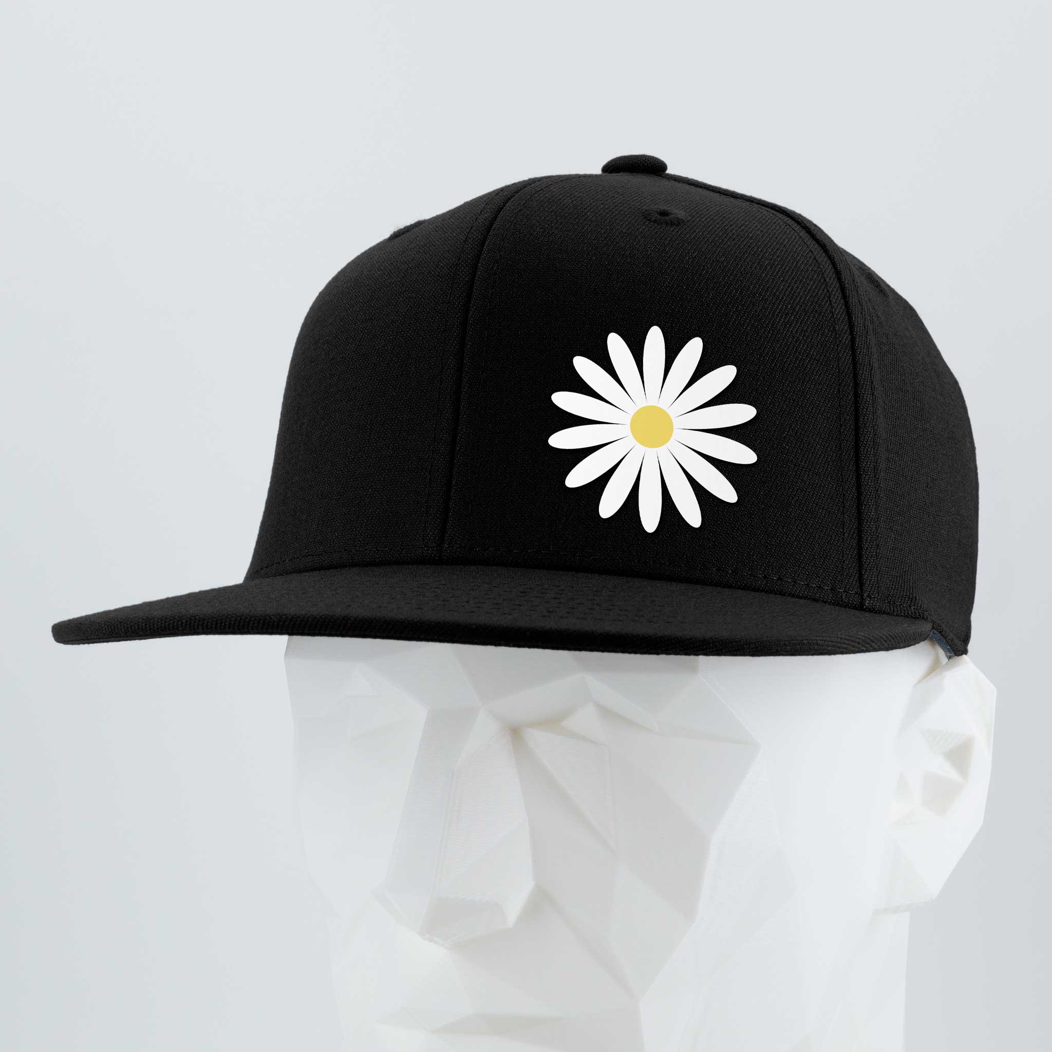 The Daisy (Silicone) by Eternal Glyph is a black Flexfit® flat bill cap with a white daisy and yellow center embroidered on the front, shown on a white geometric mannequin head against a plain light background.
