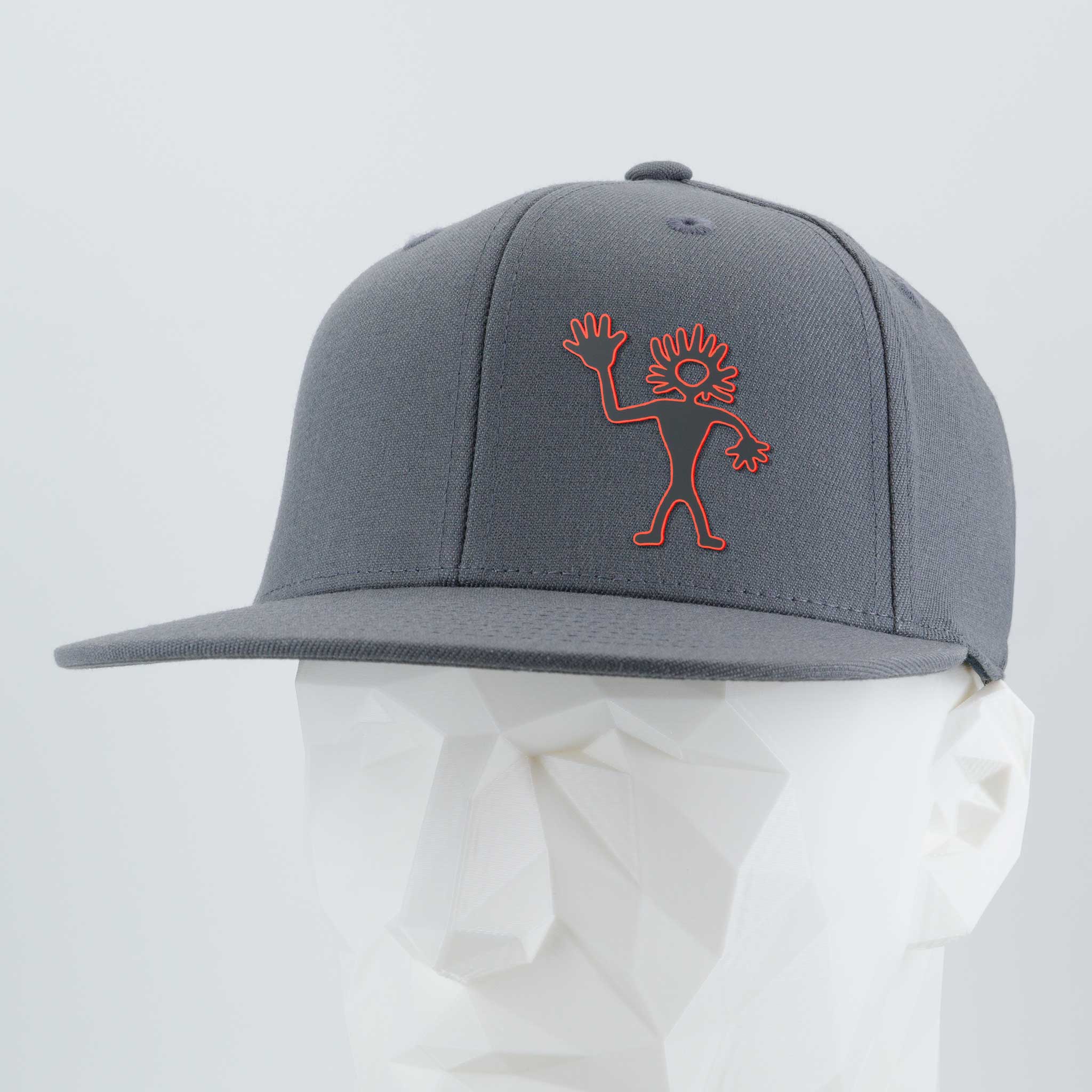 The Eternal Glyph Flexfit® Flat Bill Premium Fitted Cap in dark gray features a red outlined abstract Friendly Man Petroglyph (Silicone) on the front, inspired by Moab, Utah rock art, shown on a white mannequin against a light gray background.