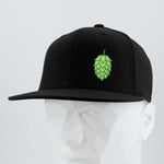 The Eternal Glyph Hop Cone (Silicone) Flexfit® Flat Bill Premium Fitted Cap in black showcases a green hop cone graphic, modeled on a white geometric mannequin head against a simple, light background.