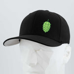 The Eternal Glyph Hop Cone (Silicone) Flexfit® Premium Wool Blend Cap is a black cap with a green hop cone symbol, shown on a white geometric mannequin head against a light gray background—ideal for beer enthusiasts.