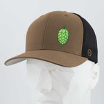 The Eternal Glyph Hop Cone (Silicone) Flexfit® Mesh Trucker Cap is a brown and black hat with a green hop cone symbol, ideal for beer lovers, shown on a faceted white mannequin head against a plain light background.