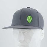 The Eternal Glyph Hop Cone (Silicone) Flexfit® Flat Bill Premium Fitted Cap in gray, featuring a green hop cone graphic, is shown on a white geometric mannequin head with a light gray background.