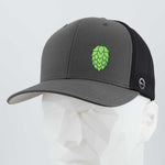 The Eternal Glyph Hop Cone (Silicone) Flexfit® Mesh Trucker Cap features a gray and black design with a green hop cone symbol, ideal for beer lovers, shown on a white geometric mannequin head.