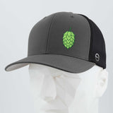 The Eternal Glyph Hop Cone (Silicone) Flexfit® Mesh Trucker Cap features a gray and black design with a green hop cone symbol, ideal for beer lovers, shown on a white geometric mannequin head.