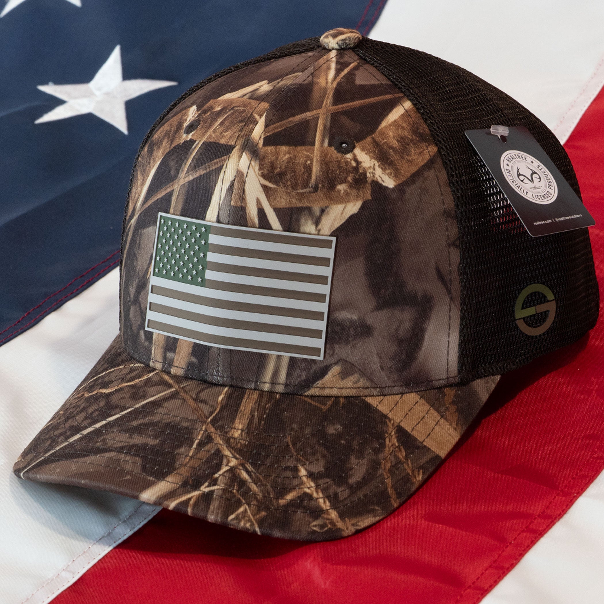 The Eternal Glyph American Flag, Realtree® Camo (Double Layer Silicone) Flexfit® Mesh Trucker Cap in Realtree® Max features a black mesh back and grayscale flag patch—an ideal outdoor cap for proud patriots.