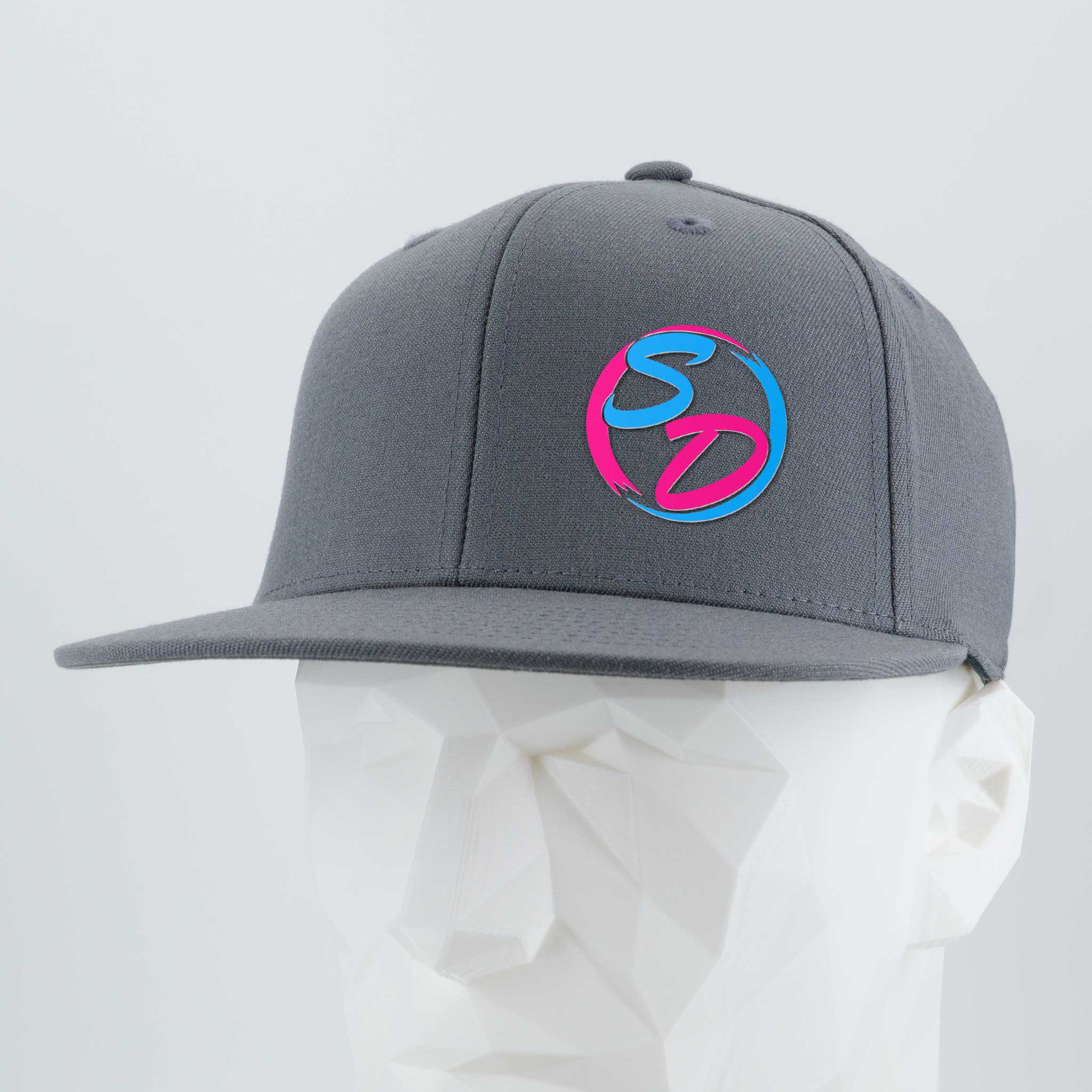 A Dark Gray Flexfit® Flat Bill Premium Fitted Cap from Eternal Glyph, featuring a blue and pink SD silicone logo on the front, is showcased on a white geometric mannequin head against a light background—an ideal choice for dedicated members of the Staying Driven online fitness crew.