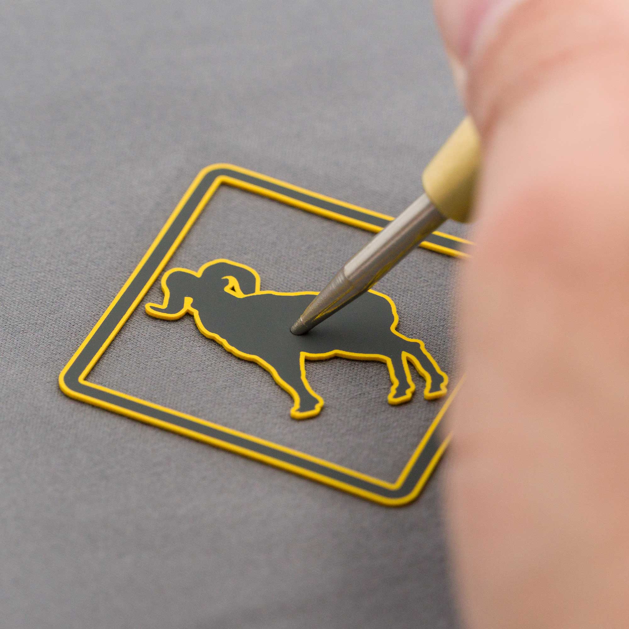 A close-up of a hand using a tool to carefully peel a sticker shaped like a ram from a square yellow outline on a gray surface.
