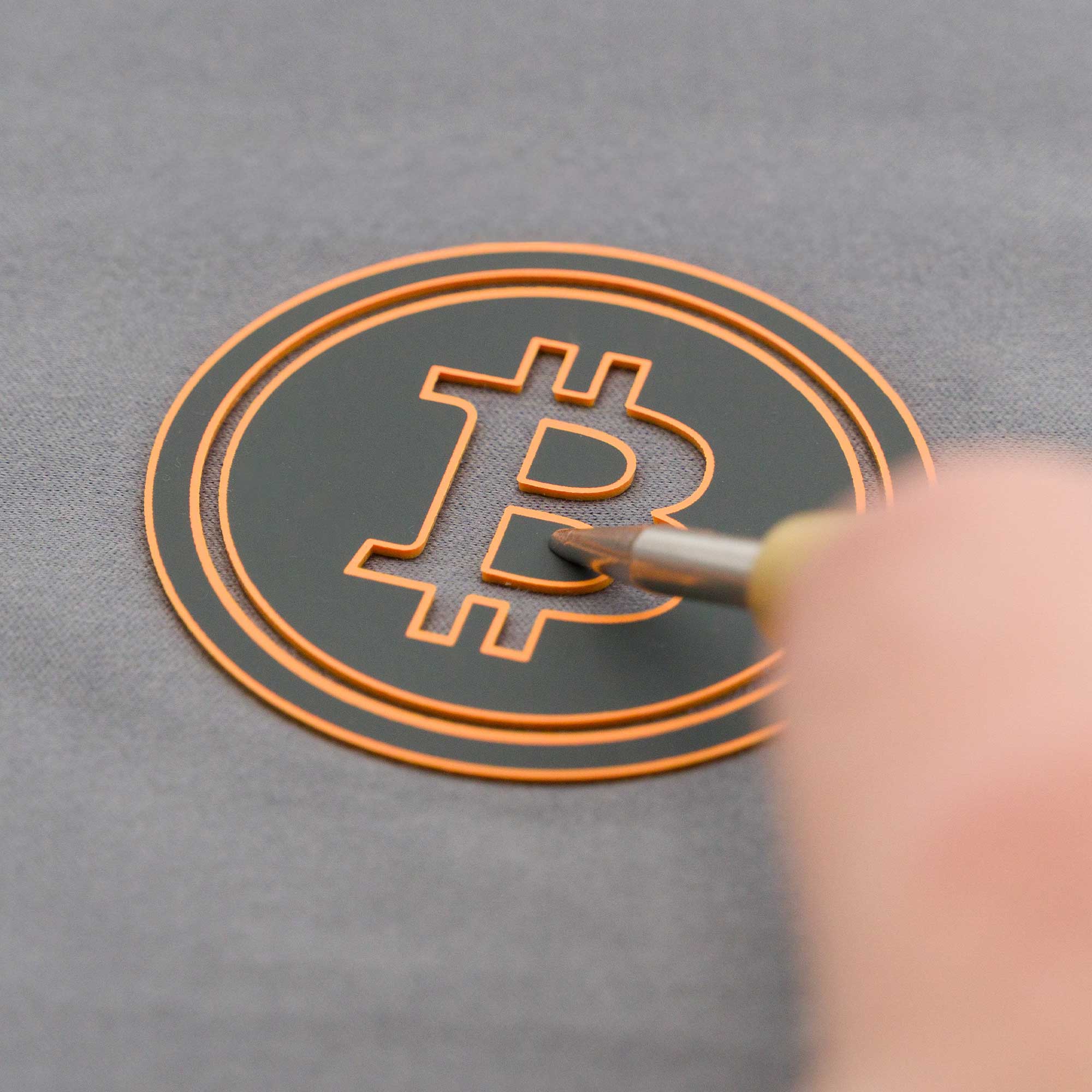 A hand uses a tool to etch or trace the bitcoin symbol, which is outlined in orange on a dark circular surface against a gray background.