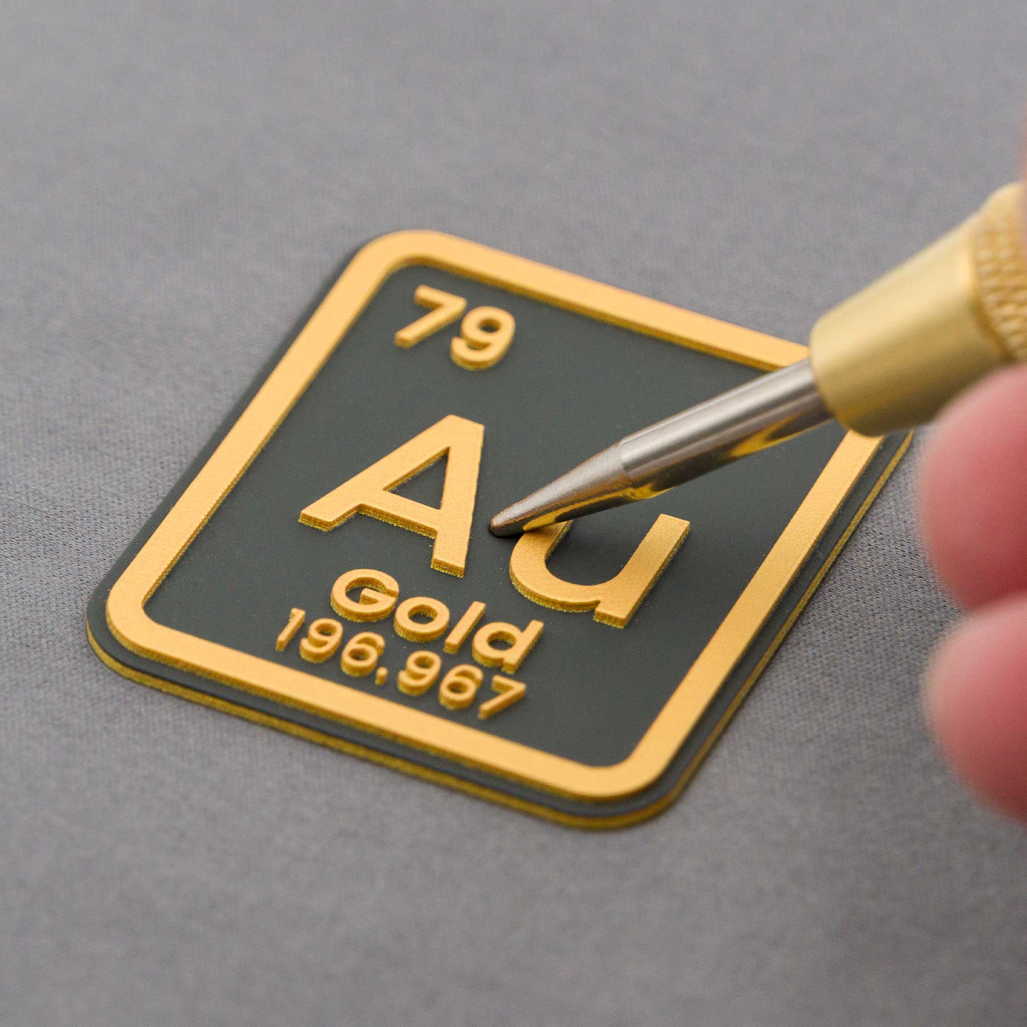 A close-up of a hand holding a stylus near a gold and black metallic plaque displaying the periodic table symbol for gold: Au, atomic number 79, and atomic mass 196.967.