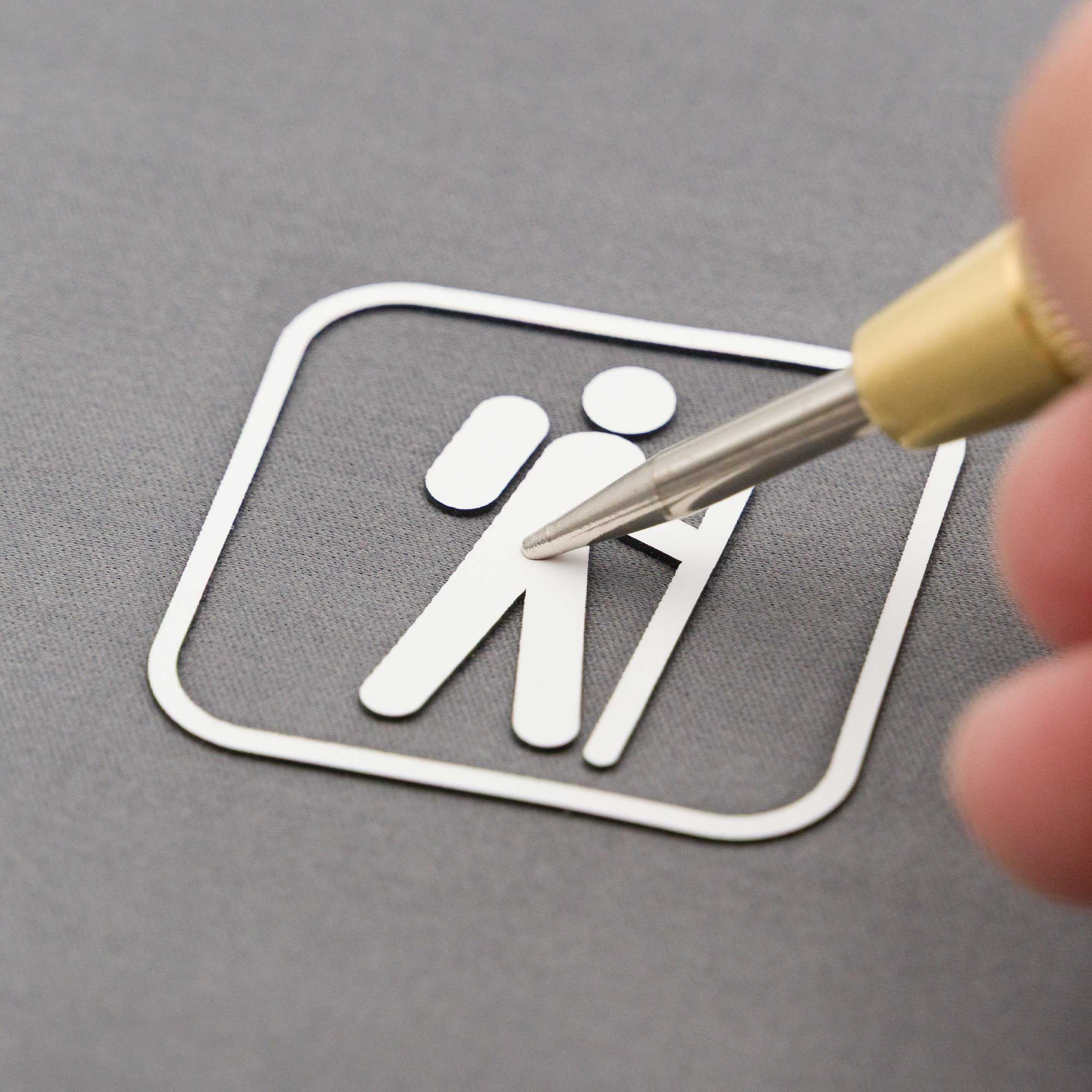 A close-up of a hand holding a precision knife, cutting out a white hiking symbol sticker on a gray surface. The symbol shows a person with a backpack and walking stick inside a rounded square.