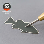 A metal tool presses onto a dark gray silicone fish patch, set on a gray surface. In the corner, the Eternal Glyph logo appears—Large Mouth Bass (Silicone) on a Flexfit® Flat Bill Premium Fitted Cap, ideal for bass fishing fans.