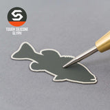 A metal tool presses onto a dark gray silicone fish patch, set on a gray surface. In the corner, the Eternal Glyph logo appears—Large Mouth Bass (Silicone) on a Flexfit® Flat Bill Premium Fitted Cap, ideal for bass fishing fans.