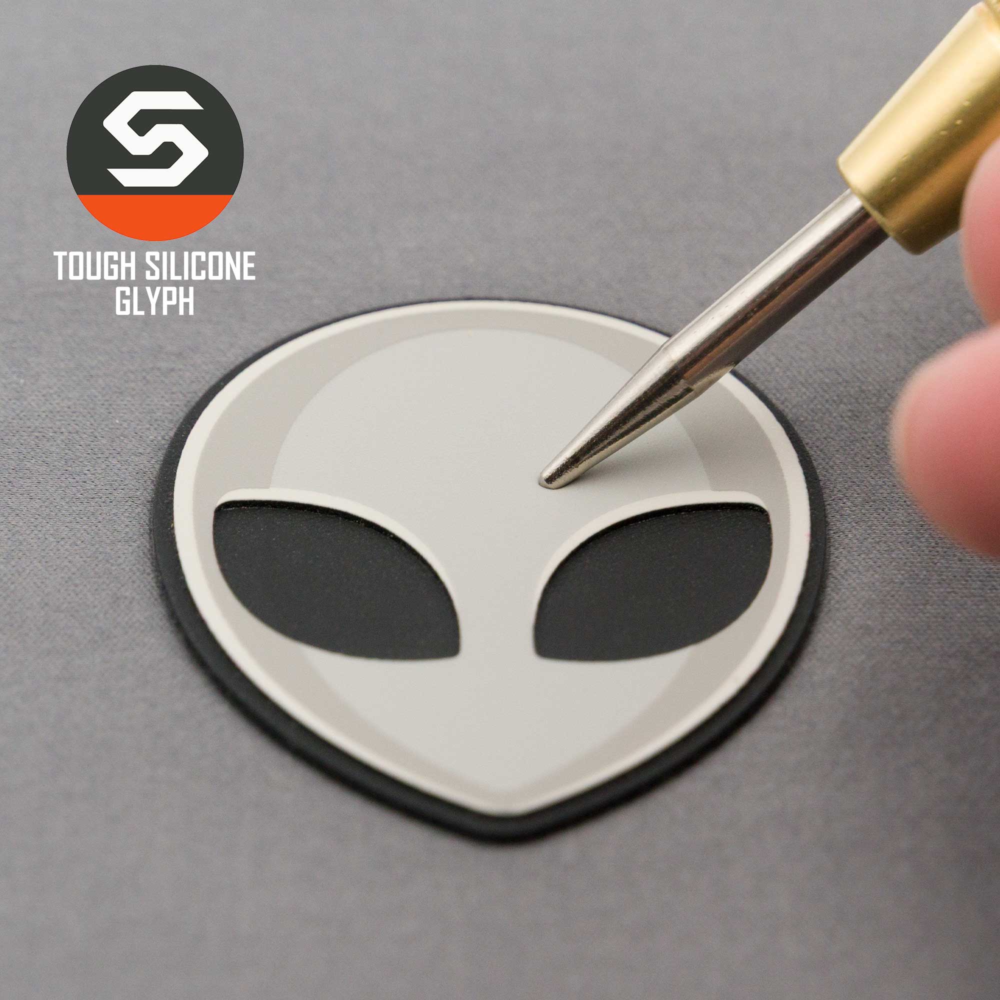 A pen tip points at an Alien Gray (Double Layer Silicone) Flexfit® Mesh Trucker Cap by Eternal Glyph, featuring a gray alien head patch with large black eyes and a stylized S logo, channeling pop culture UFO vibes.