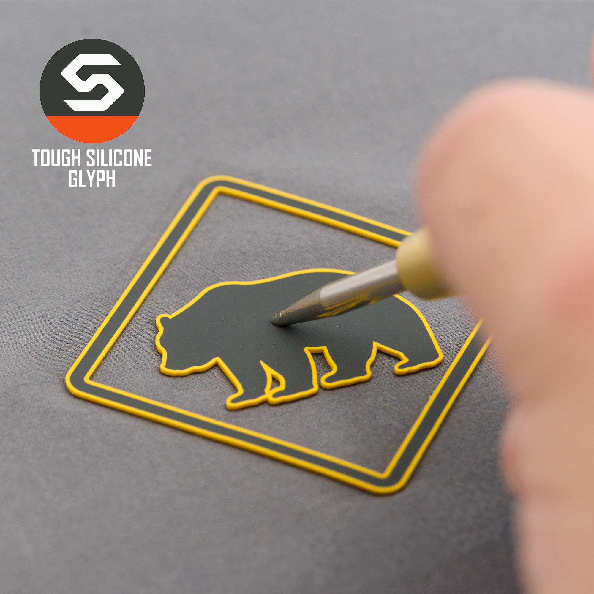 A hand traces the outline of a bear-shaped patch with a yellow border and bold bear symbol on a gray surface. "Bear Crossing Sign (Silicone) - Flexfit® Mesh Trucker Cap" by Eternal Glyph appears in the top-left corner.