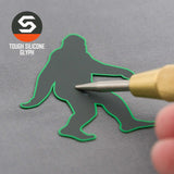A hand points a tool at a gray Flexfit® Premium Wool Blend Cap featuring Eternal Glyph’s Bigfoot Silhouette in green outline. The top left shows the Eternal Glyph logo and highlights this unique adventure icon in tough silicone.