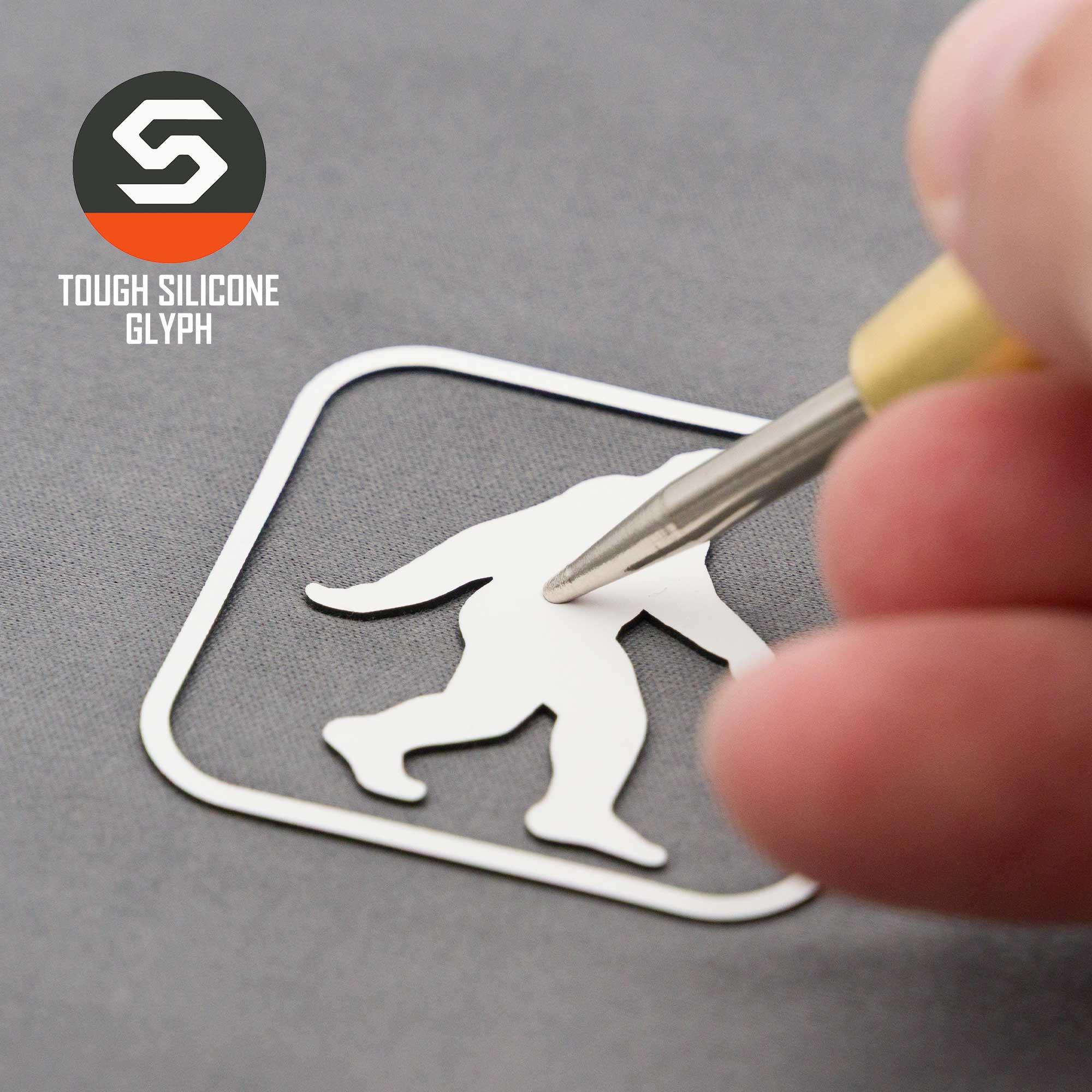 A hand peels a white Bigfoot silhouette sticker from its backing, evoking cryptid folklore. The text "Bigfoot Trail Sign (Silicone) - Flexfit® Mesh Trucker Cap" by Eternal Glyph appears next to a stylized “S” logo on gray.