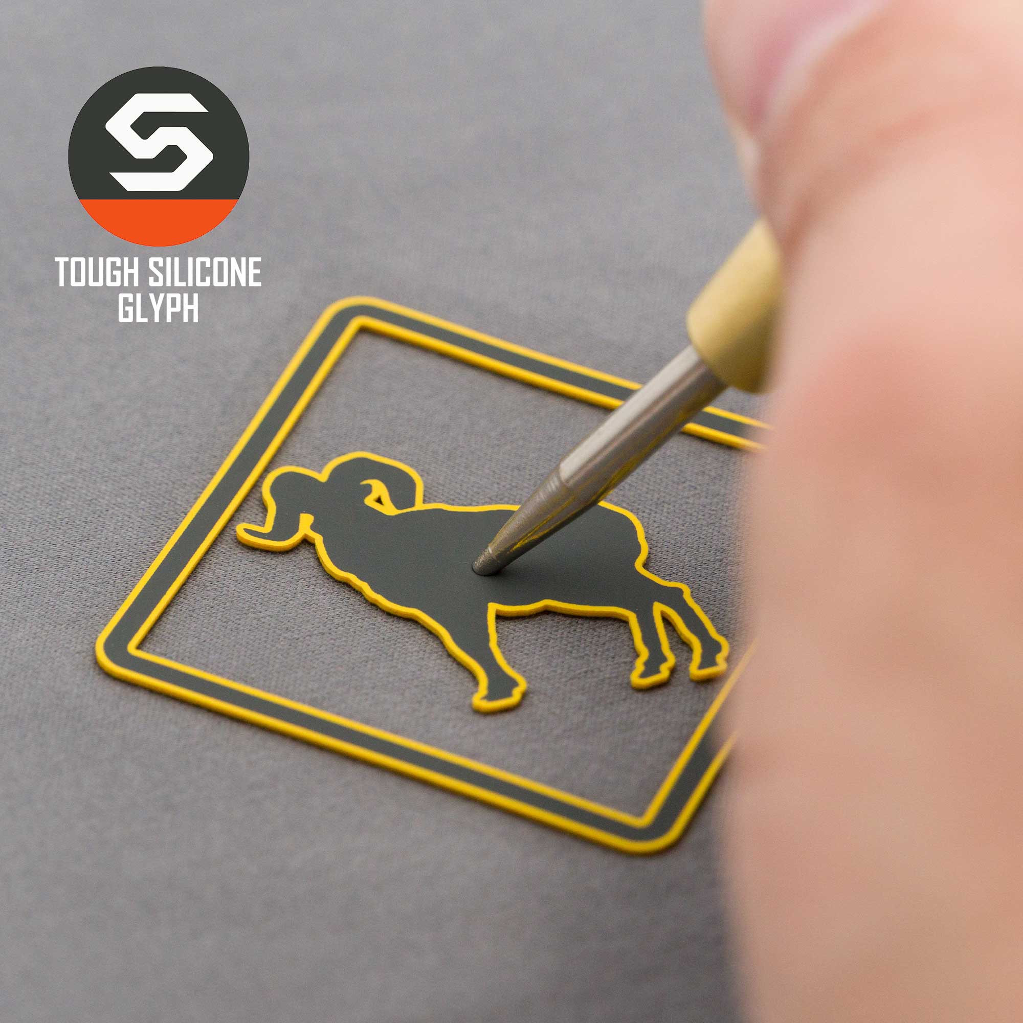 A hand uses a stylus to touch a yellow-edged silicone Bighorn Sheep Crossing Sign glyph inside a square. The gray background features the Eternal Glyph logo with "TOUGH SILICONE GLYPH" at top left. Product: Flexfit® Mesh Trucker Cap.