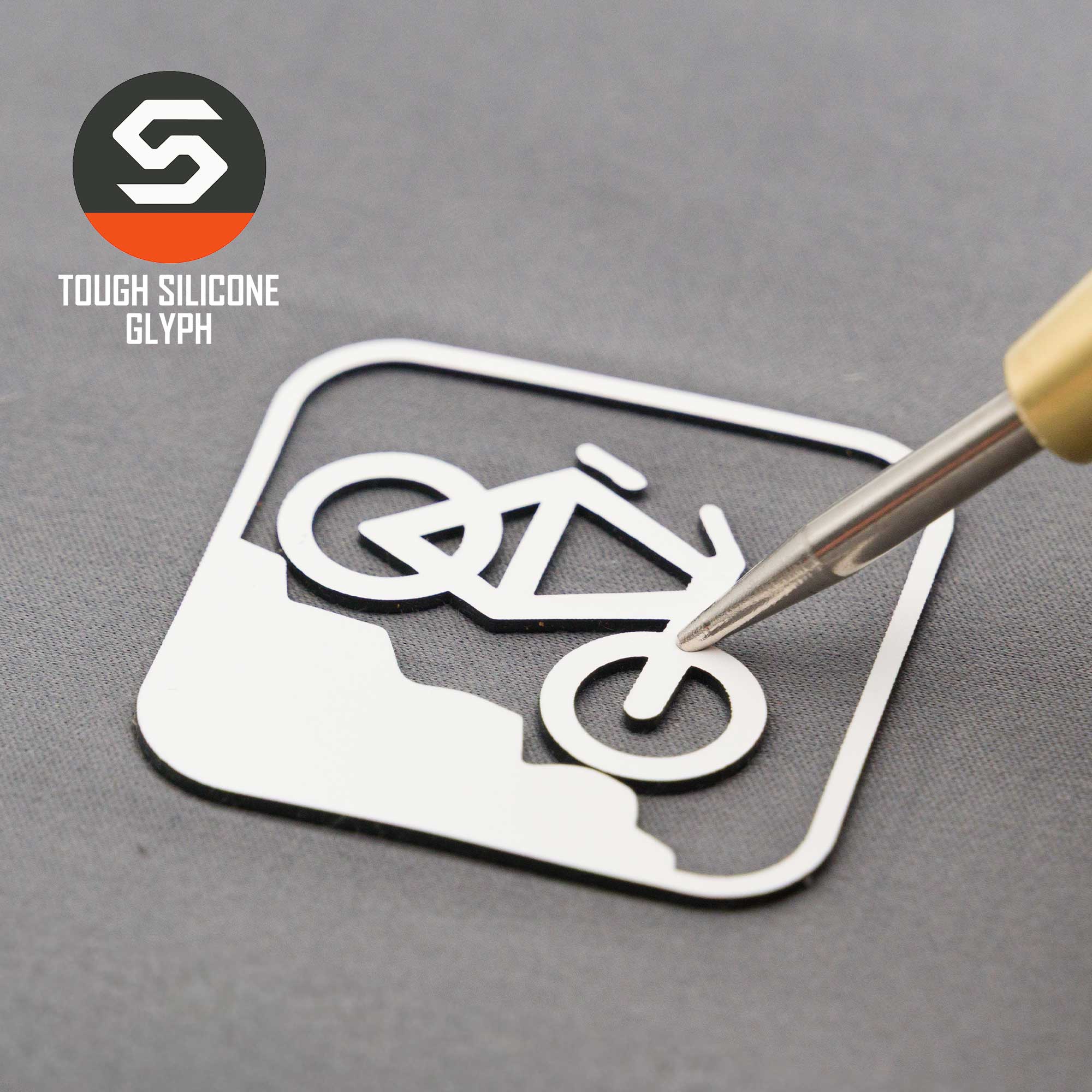A metal tool lifts a white sticker with a mountain bike downhill icon on gray. Top left: Eternal Glyph hexagonal logo and "Mountain Bike Downhill (Silicone) - Flexfit® Mesh Trucker Cap" appear.