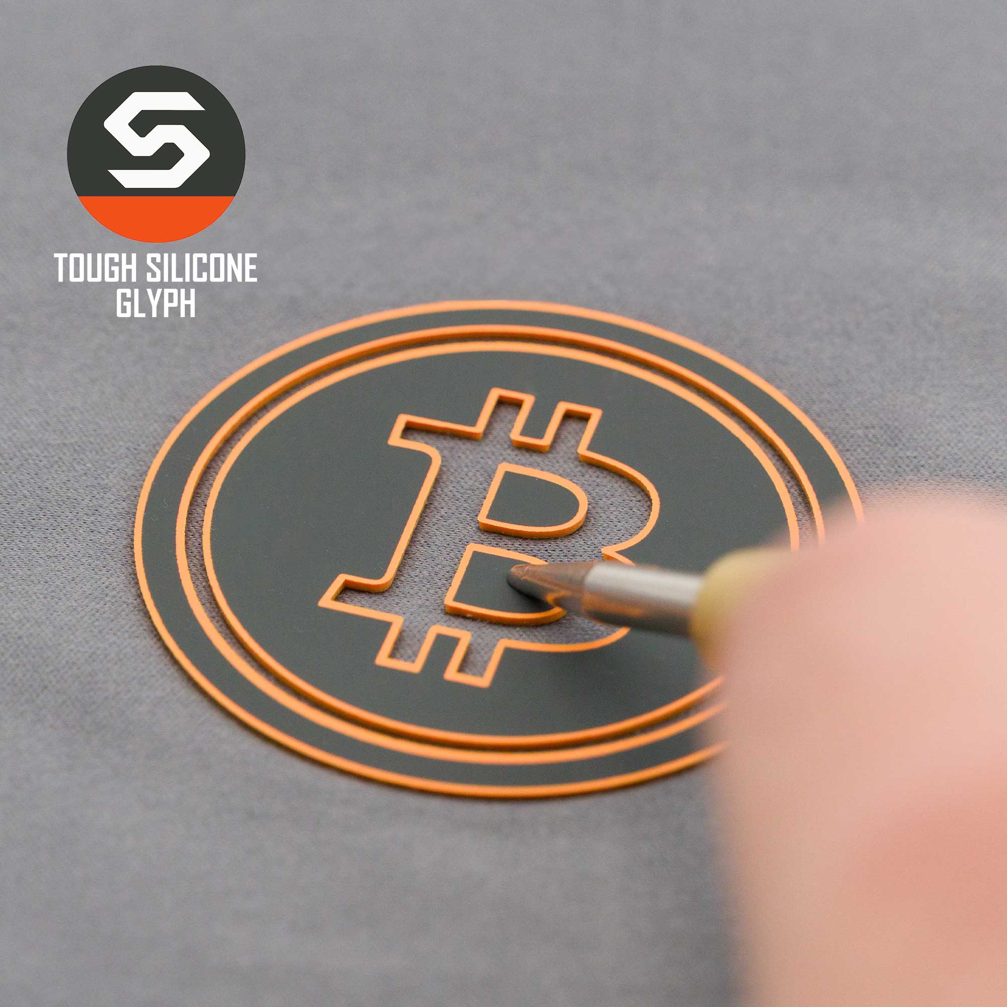 A hand peels a black-and-orange round sticker with the Bitcoin symbol, referencing cryptocurrency. Top left shows Eternal Glyph's logo and text: "Bitcoin Silhouette (Silicone) - Flexfit® Mesh Trucker Cap.