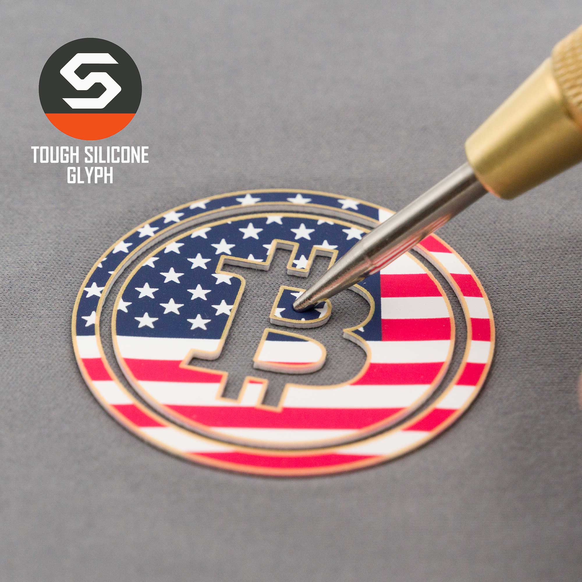 A tool engraves a Bitcoin U.S.A. glyph on the Flexfit® Cool & Dry Cap by Eternal Glyph, featuring an American flag design to symbolize financial freedom. "Tough Silicone Glyph" and the stylized S logo appear in the upper left corner.