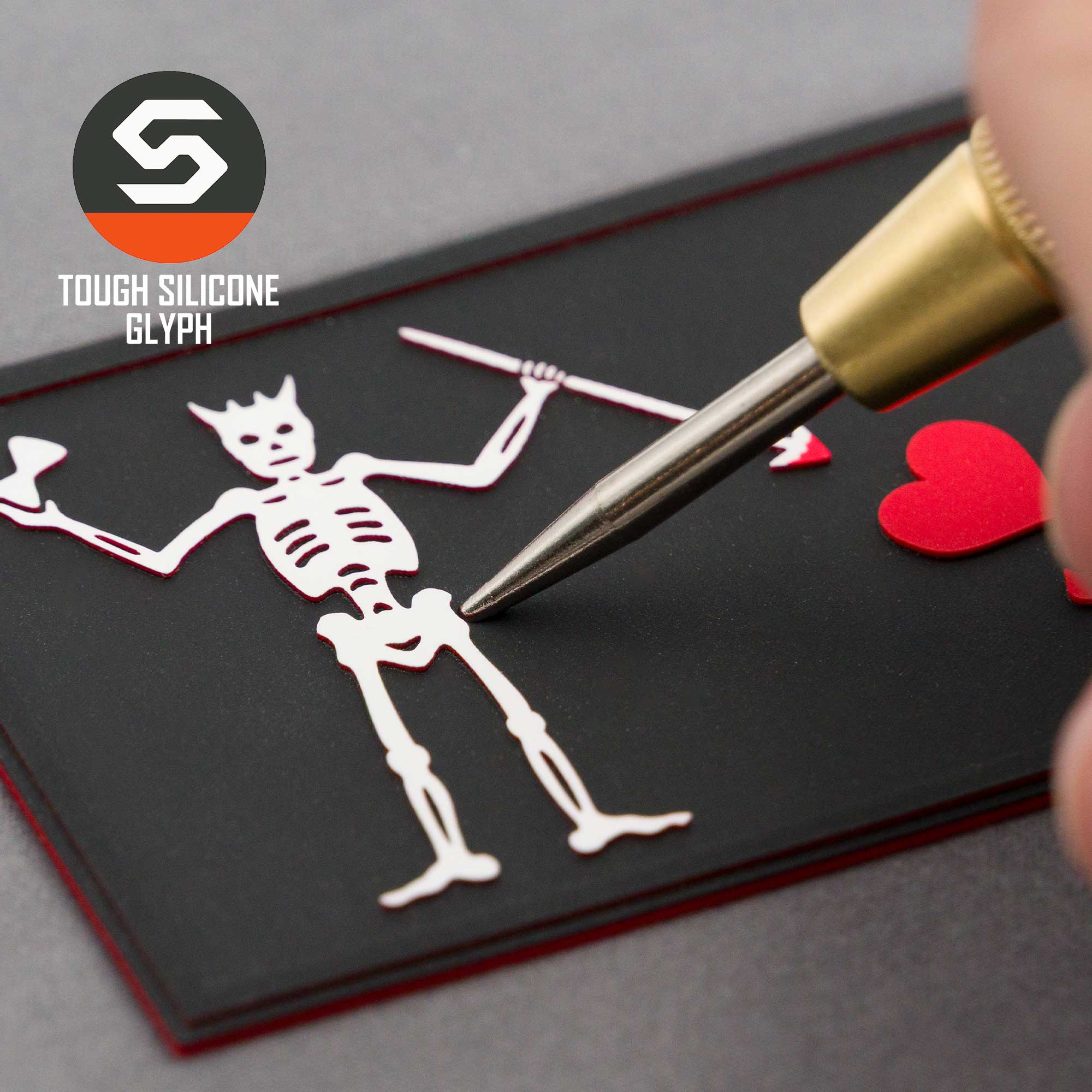 A close-up of a hand working on a Black Beard Pirate Flag (Double Layer Silicone) - Flexfit® Cool & Dry Cap by Eternal Glyph, featuring a white skeleton and red hearts on black, with the Tough Silicone Glyph logo in the top left corner.