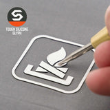 A hand uses a stylus to trace the white Campfire Trail Sign (Silicone) icon, featuring logs and flame within a square, on a gray surface. The Flexfit® Mesh Trucker Cap by Eternal Glyph is branded at the top left.