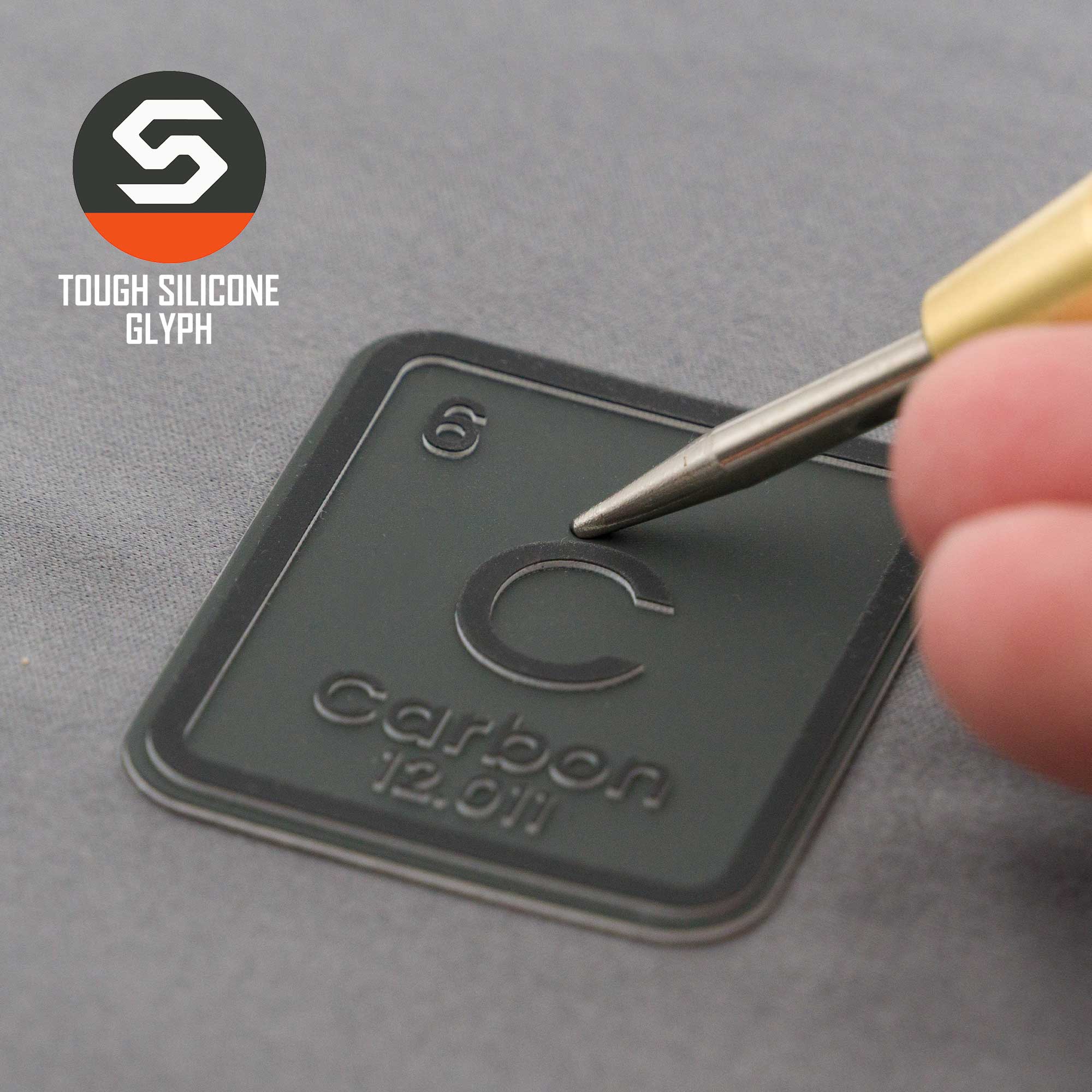 A hand presses an Eternal Glyph Carbon Element (Double Layer Silicone) patch—shaped like the carbon periodic table tile, labeled C, Carbon, 12.01—onto a gray Flexfit® Mesh Trucker Cap. "Tough Silicone Glyph" appears next to the logo.