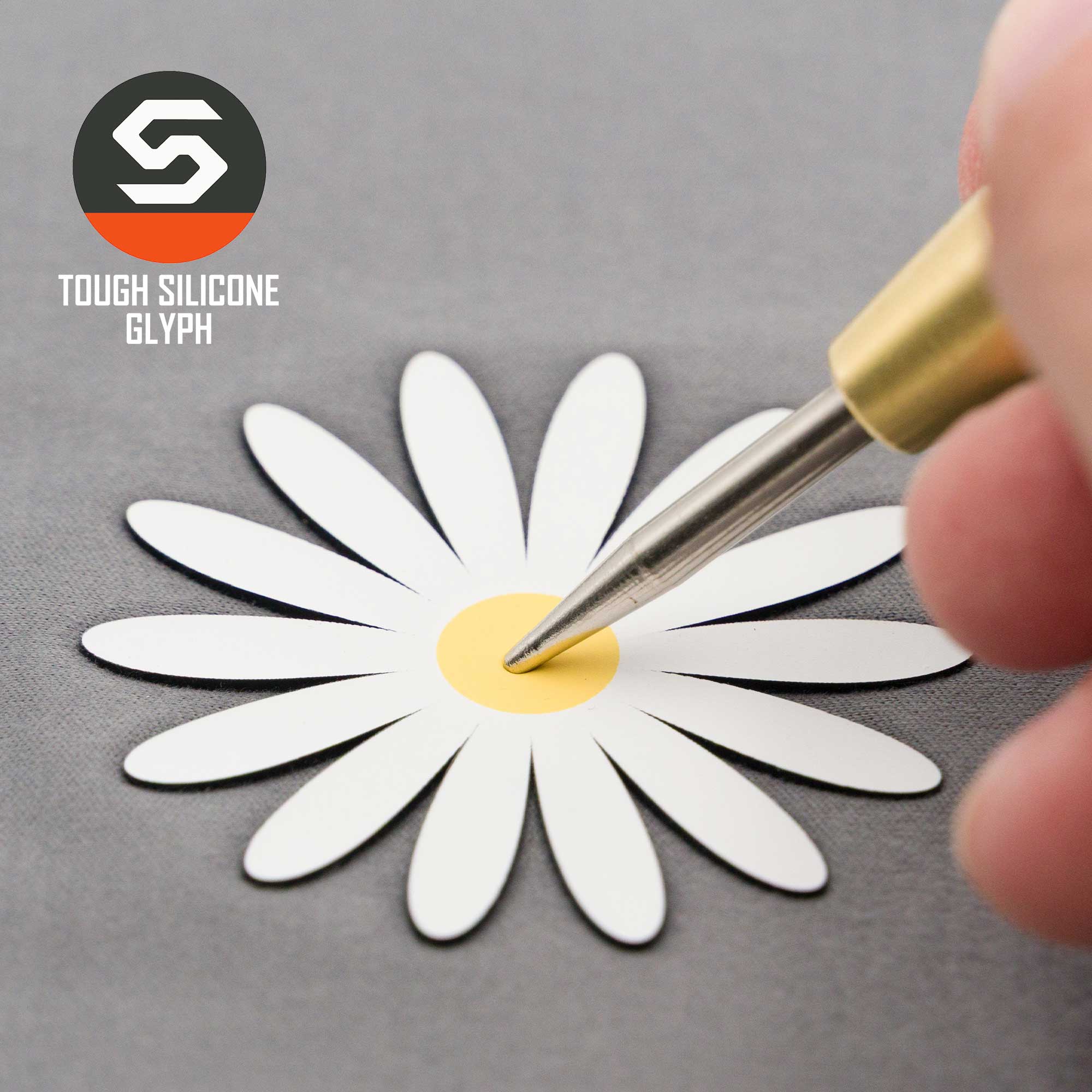 A hand holding a precision tool touches the yellow center of a white daisy—an optimism symbol—on the gray Flexfit® Mesh Trucker Cap, The Daisy (Silicone) by Eternal Glyph, with its logo in the corner.
