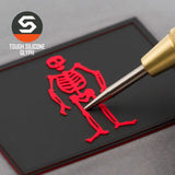 A gold tool points to a red skeletal figure on a black card, evoking the Edward Low Pirate Flag. The Eternal Glyph logo and "Tough Silicone Glyph" text appear, resembling a pirate flag glyph on a Flexfit® Cool & Dry Cap.