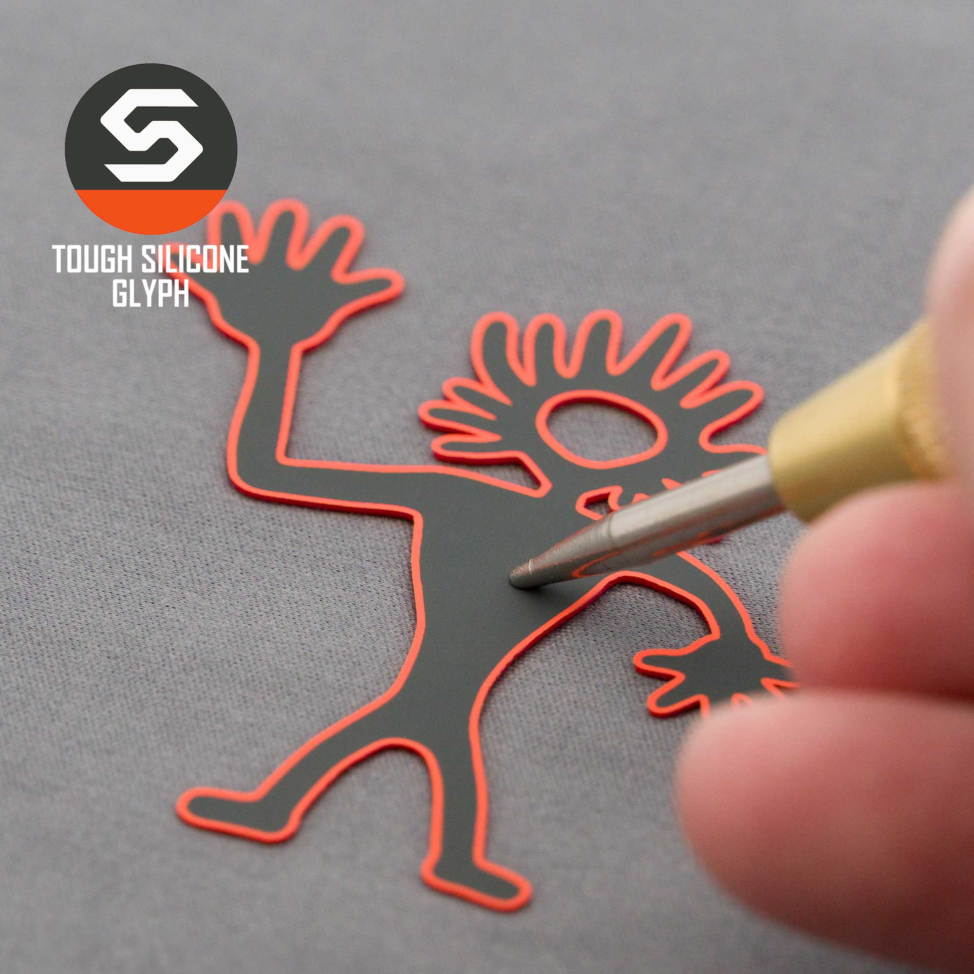 A hand uses a tool to press on a black and red outlined silicone figure with spiky hair and raised arms, inspired by the Friendly Man Petroglyph from Moab, Utah. Eternal Glyph’s Flexfit® Mesh Trucker Cap logo appears in the corner.