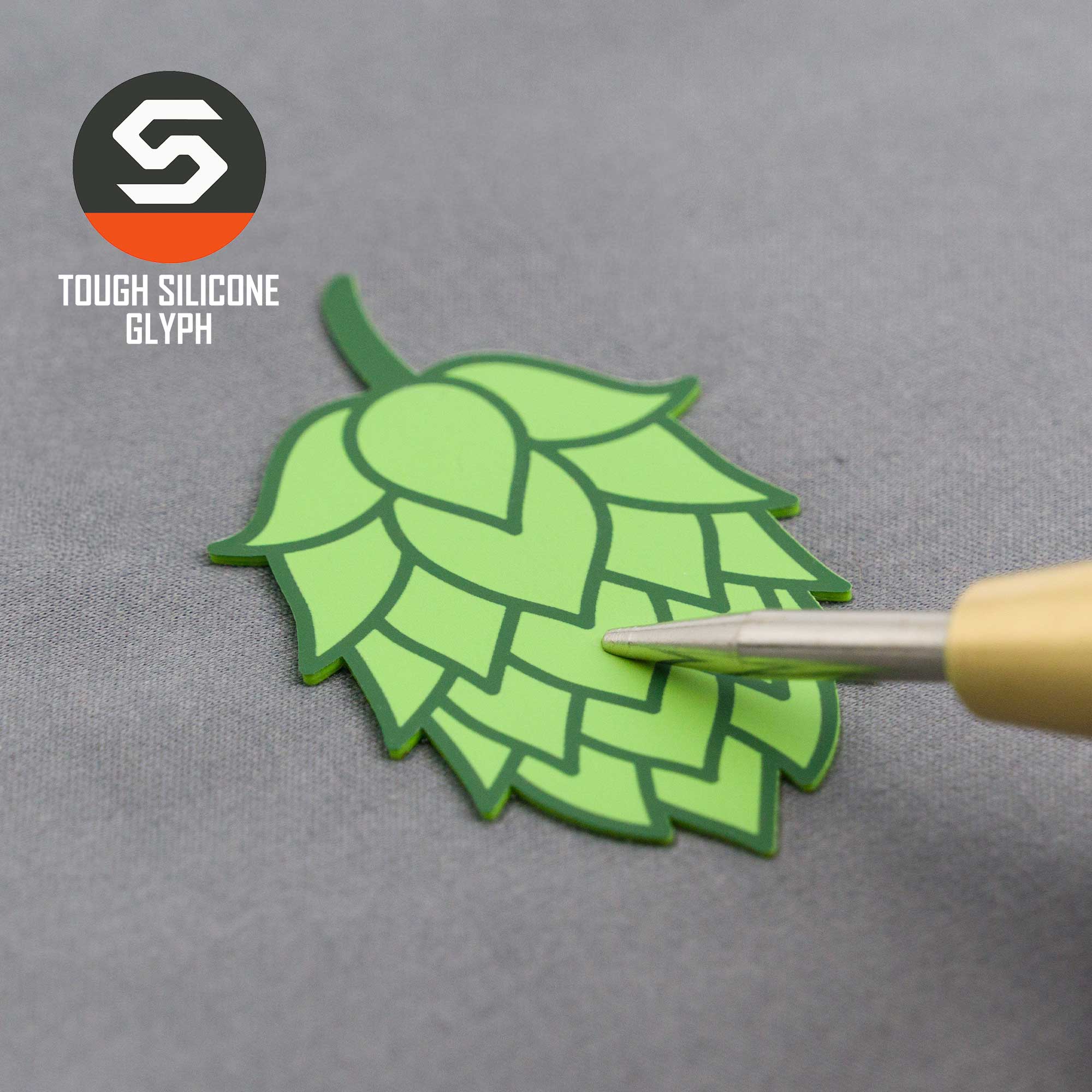 A pen points to a green hop cone-shaped silicone patch on a gray surface. In the top left, "Eternal Glyph" and an S logo appear—this detail is from the Hop Cone (Silicone) Flexfit® Mesh Trucker Cap, perfect for beer lovers.
