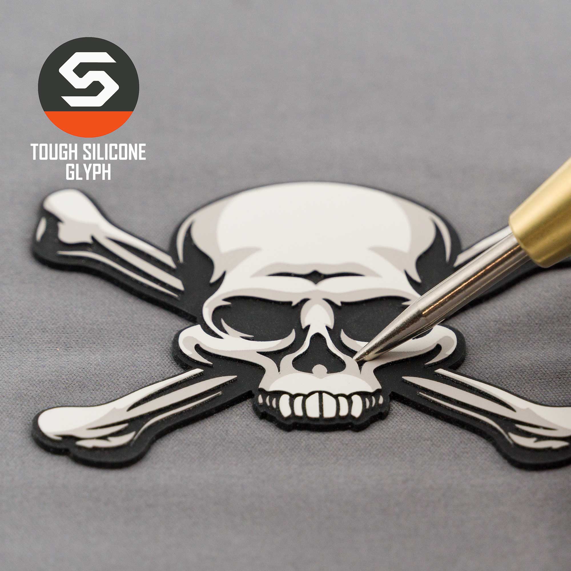 A gold pen points at a black and white silicone Pirate Skull & Crossbones patch by Eternal Glyph on gray fabric. “TOUGH SILICONE GLYPH” appears near the logo, accenting this modern Jolly Roger on a Flexfit® Cool & Dry Cap.