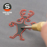 A tool presses a red and black Moab Man Petroglyph (Silicone) - Flexfit® Mesh Trucker Cap by Eternal Glyph, shaped like a cartoon creature with antlers, against a gray background. The logo and "Tough Silicone Glyph" are in the top left corner.