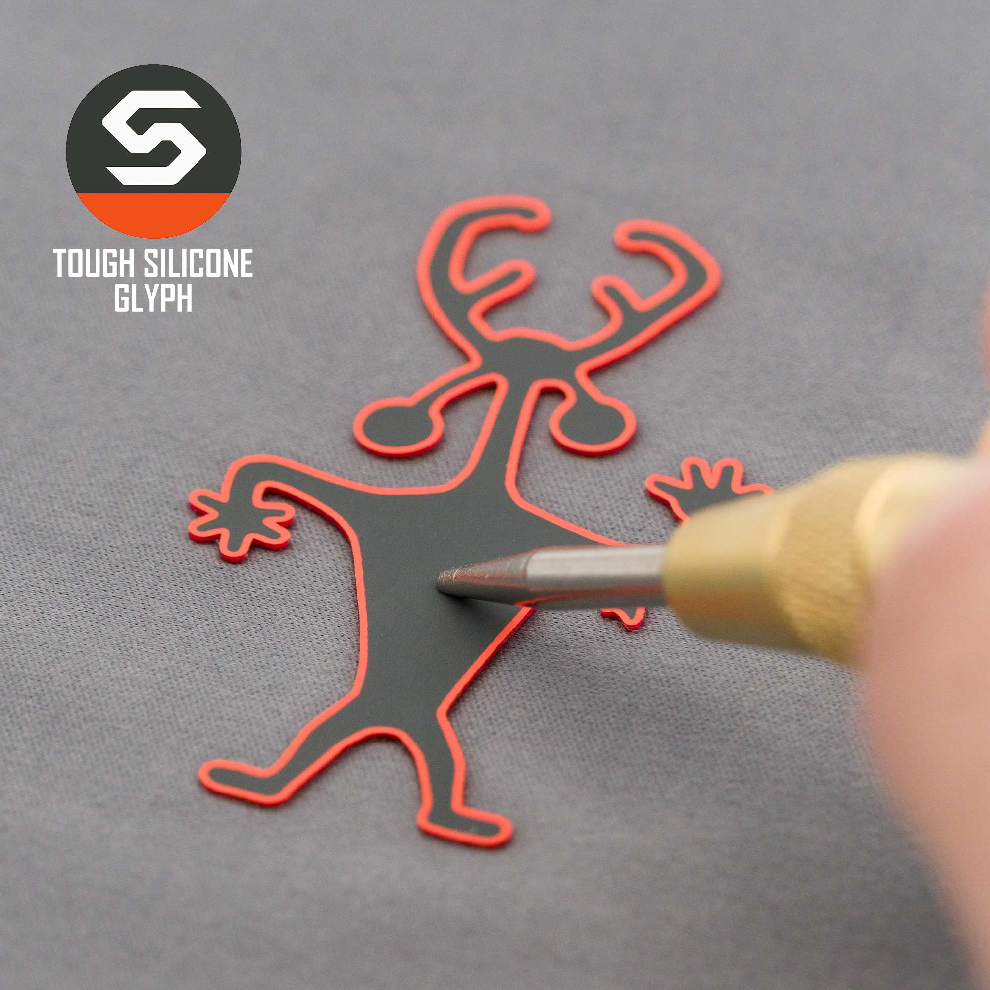 A tool presses a red and black Moab Man Petroglyph (Silicone) - Flexfit® Mesh Trucker Cap by Eternal Glyph, shaped like a cartoon creature with antlers, against a gray background. The logo and "Tough Silicone Glyph" are in the top left corner.