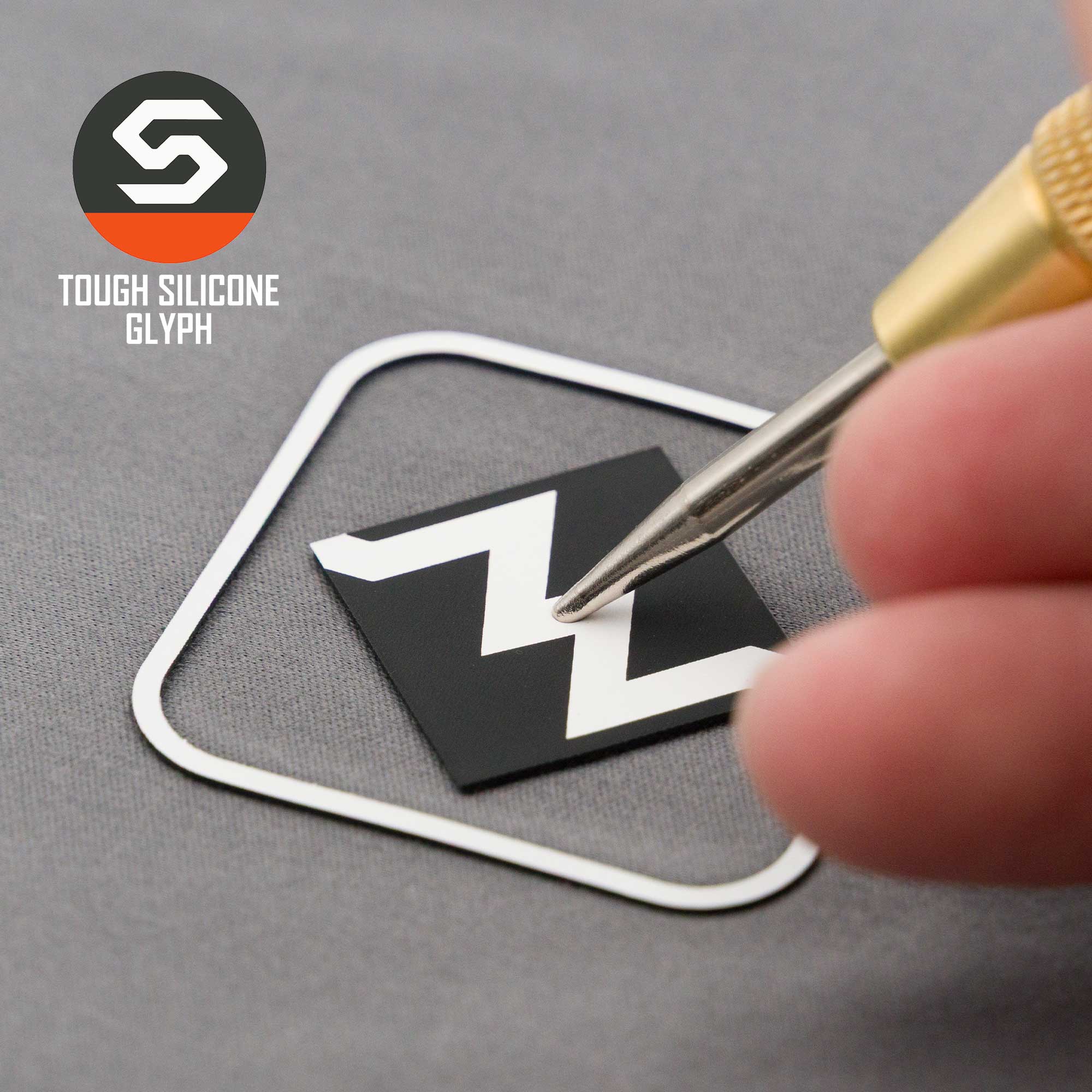 A hand presses a black and white zigzag-patterned silicone emblem, inspired by the Most Difficult Trail Sign, onto a gray cap. Text reads: "Eternal Glyph" above "TOUGH SILICONE GLYPH." Product: Flexfit® Mesh Trucker Cap.