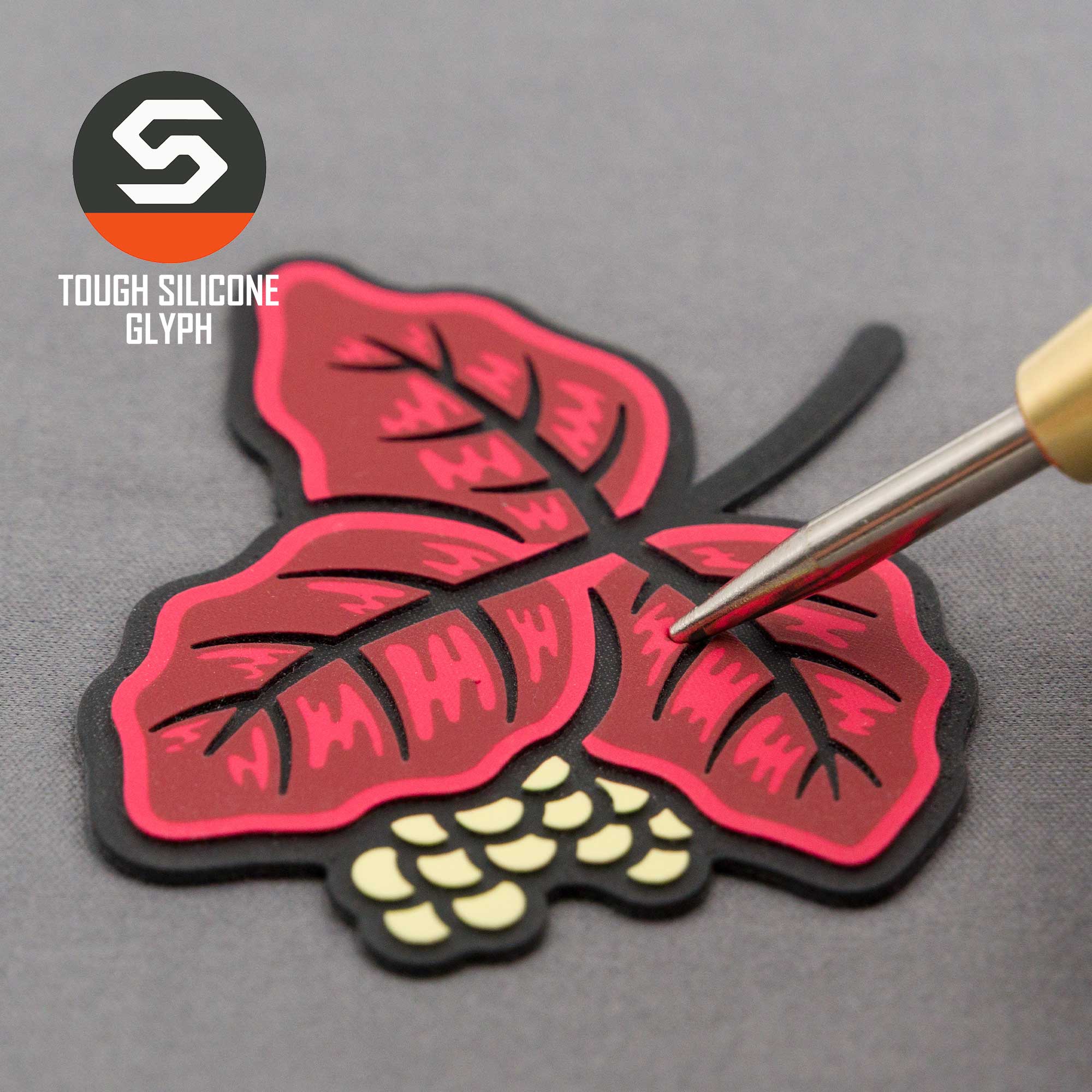 The Eternal Glyph Poison Oak (Double Layer Silicone) Flexfit® Flat Bill Premium Fitted Cap, featuring red and black poison oak leaves and berries, is displayed on a gray surface with a metallic tool beside it and the TOUGH SILICONE GLYPH logo visible.