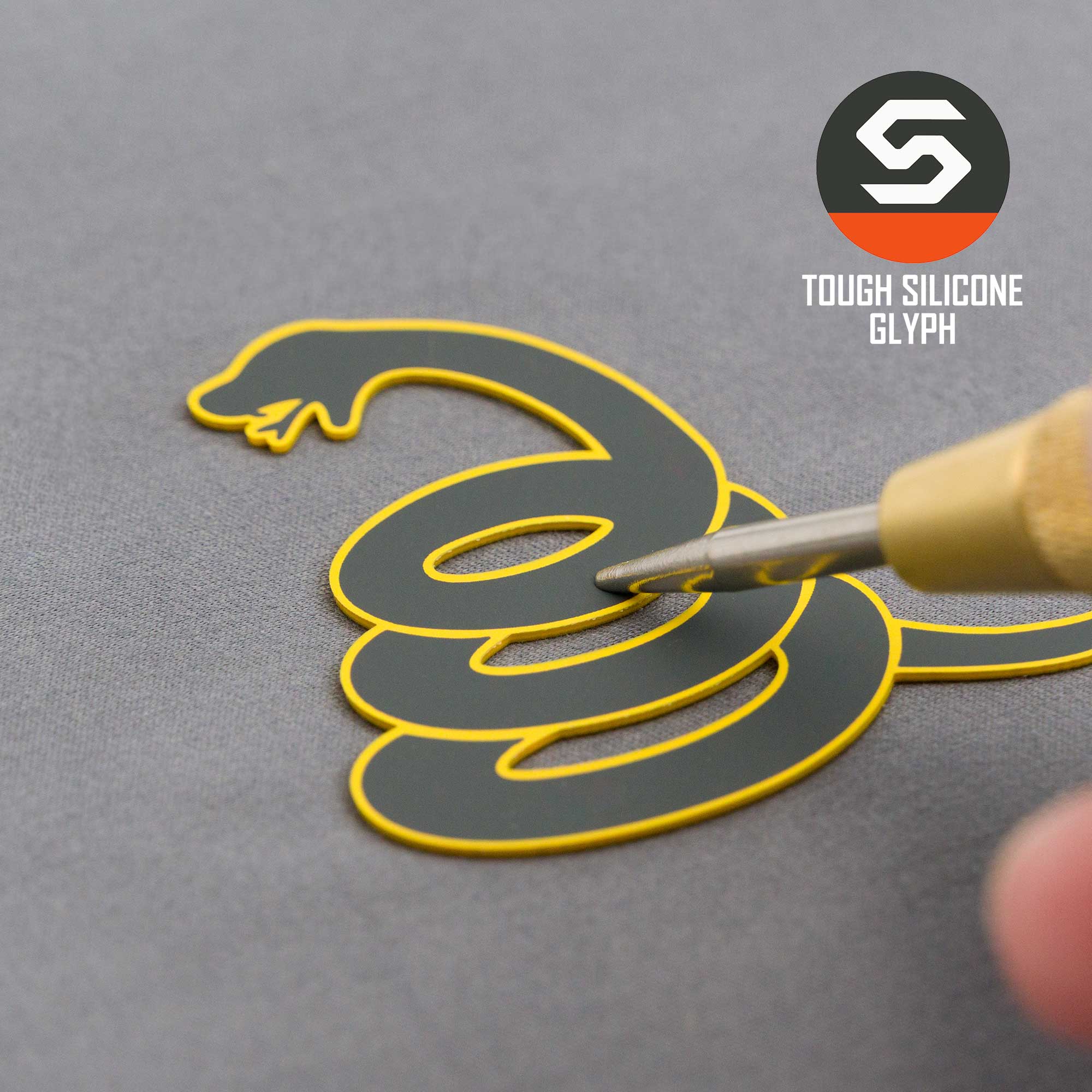 A hand uses a tool to touch the black and yellow Coiled Rattlesnake Silhouette (Silicone) on a gray Flexfit® Mesh Trucker Cap by Eternal Glyph, featuring TOUGH SILICONE GLYPH—a nod to the Gadsden flag’s symbolism of strength.