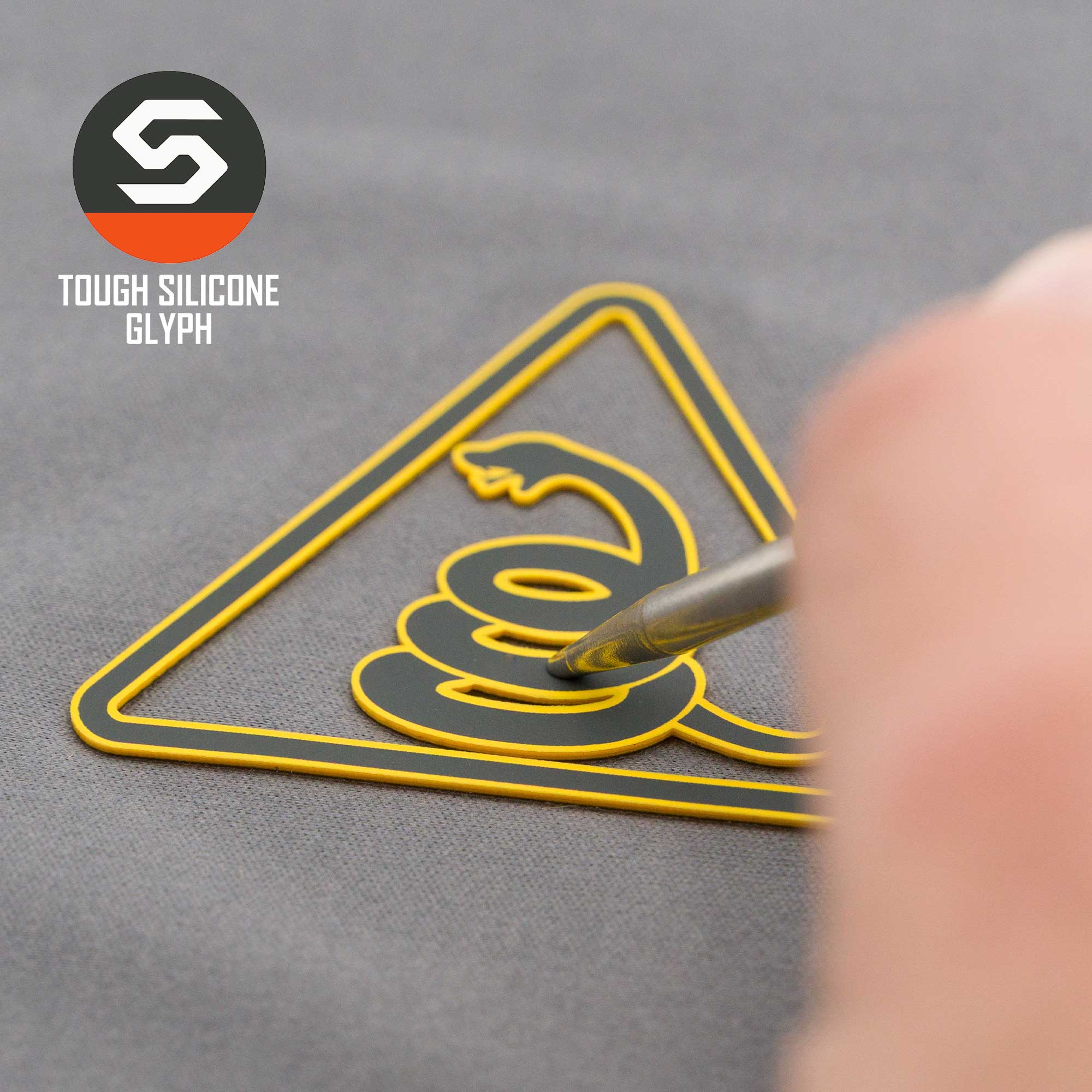 A close-up of Eternal Glyph’s Rattlesnake Warning Sign (Silicone) - Flexfit® Mesh Trucker Cap shows a yellow snake patch pressed by a tool, with the Tough Silicone Glyph logo visible on the gray background.