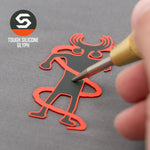 A hand uses a tool on a red and black silicone Moab Shaman Petroglyph by Eternal Glyph, shaped like a horned human and set on a gray surface. "TOUGH SILICONE GLYPH" appears next to the logo. Product: Flexfit® Mesh Trucker Cap.