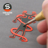 A hand uses a tool on a red and black silicone Moab Shaman Petroglyph by Eternal Glyph, shaped like a horned human and set on a gray surface. "TOUGH SILICONE GLYPH" appears next to the logo. Product: Flexfit® Mesh Trucker Cap.