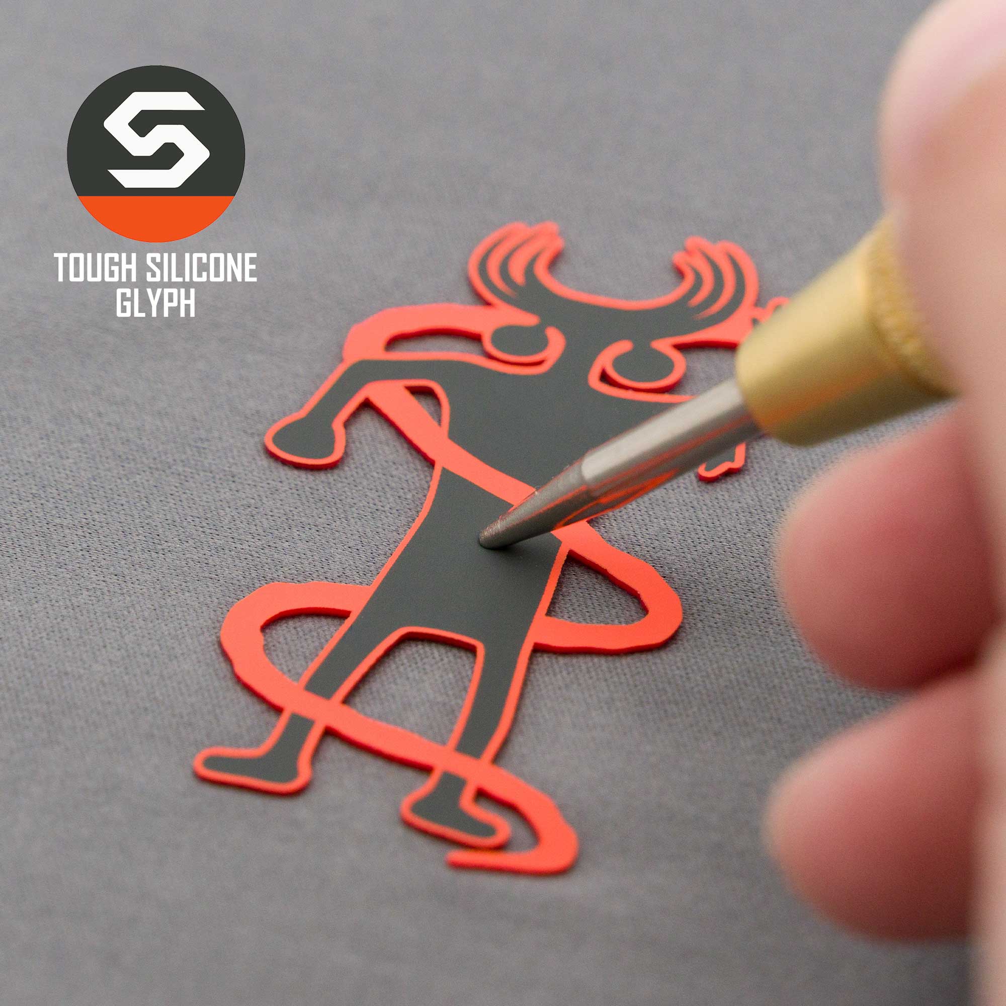A hand uses a tool on a red and black silicone Moab Shaman Petroglyph by Eternal Glyph, shaped like a horned human and set on a gray surface. "TOUGH SILICONE GLYPH" appears next to the logo. Product: Flexfit® Mesh Trucker Cap.