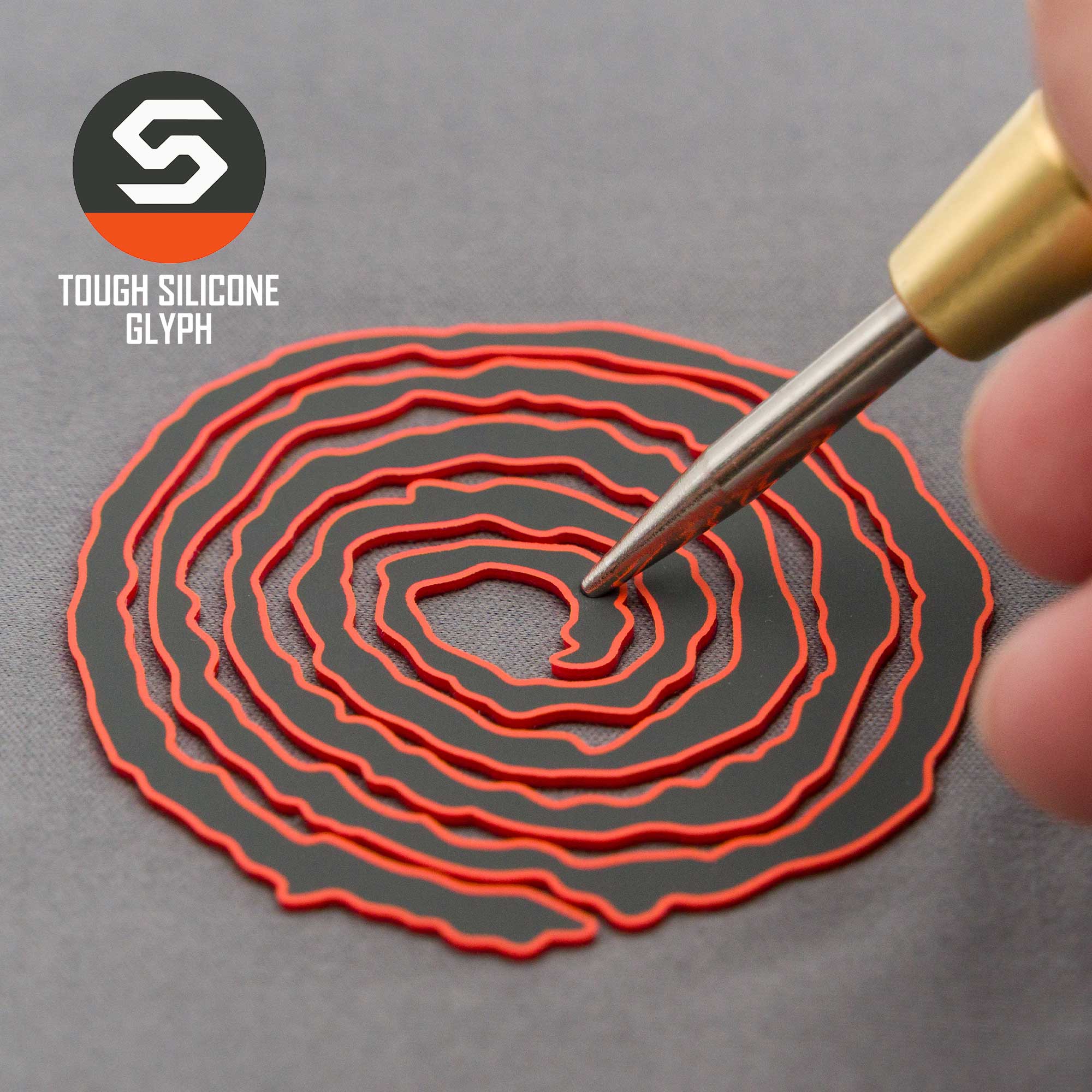 A hand holds a tool near the black and red spiral Moab Spiral Petroglyph (Silicone) on an Eternal Glyph Flexfit® Mesh Trucker Cap; "TOUGH SILICONE GLYPH" and the stylized S logo appear in the upper left corner.