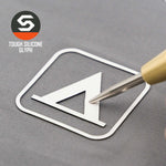 A metallic pen tip touches a white geometric glyph inside a square on gray, evoking a camping trail sign. The top left shows the Eternal Glyph logo and “Tent Camping Trail Sign (Silicone) - Flexfit® Mesh Trucker Cap.”.