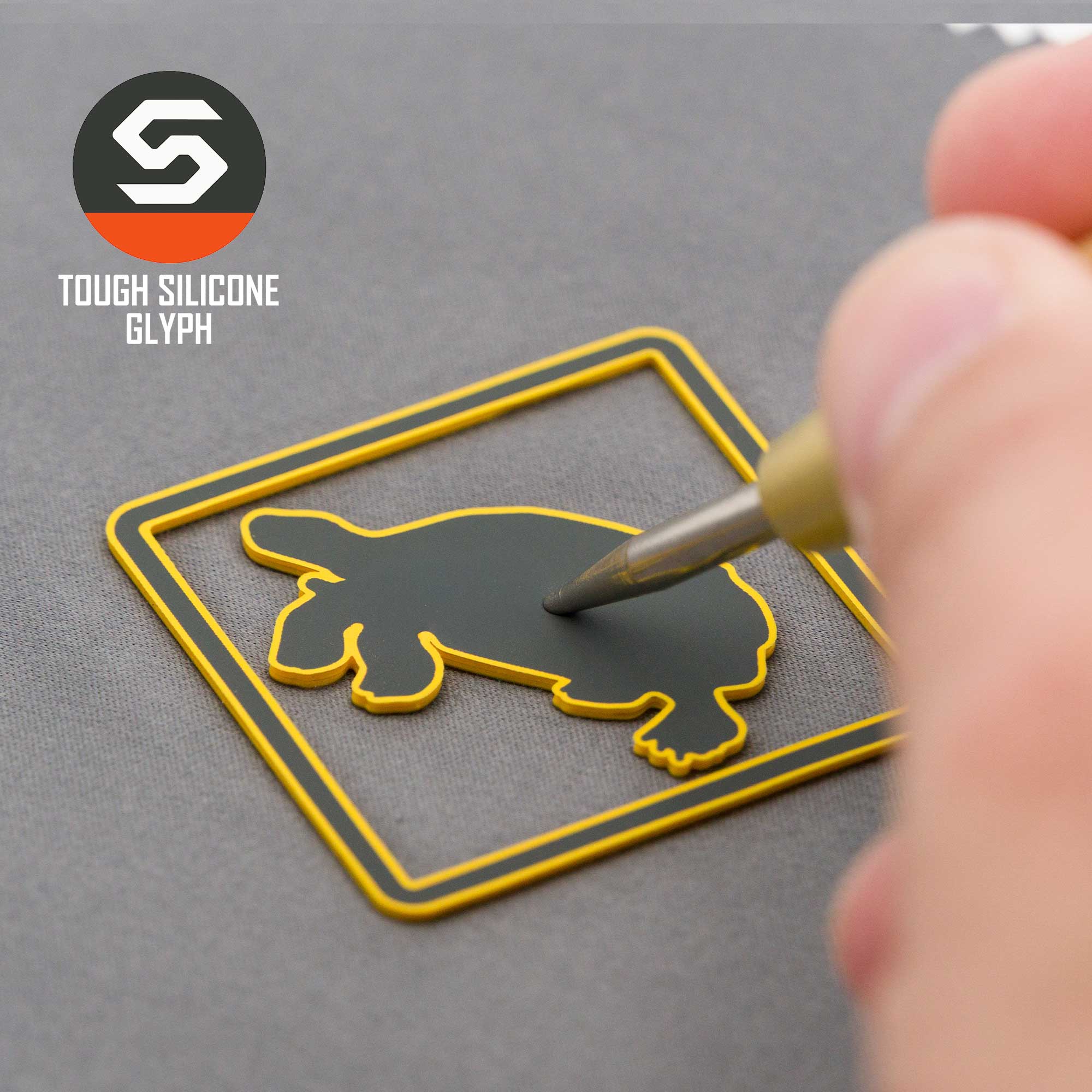 A hand applies a black platypus-shaped silicone patch inside a yellow square on the Eternal Glyph Tortoise Crossing Sign (Silicone) Flexfit® Mesh Trucker Cap, evoking resilience, with logo and TOUGH SILICONE GLYPH text in the corner.