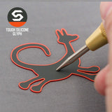 A hand presses a tool onto a flat, cat-shaped silicone patch with a red outline on a gray surface. The patch is the Eternal Glyph Moab Trickster Petroglyph (Silicone) for the Flexfit® Mesh Trucker Cap, inspired by Moab Utah designs.