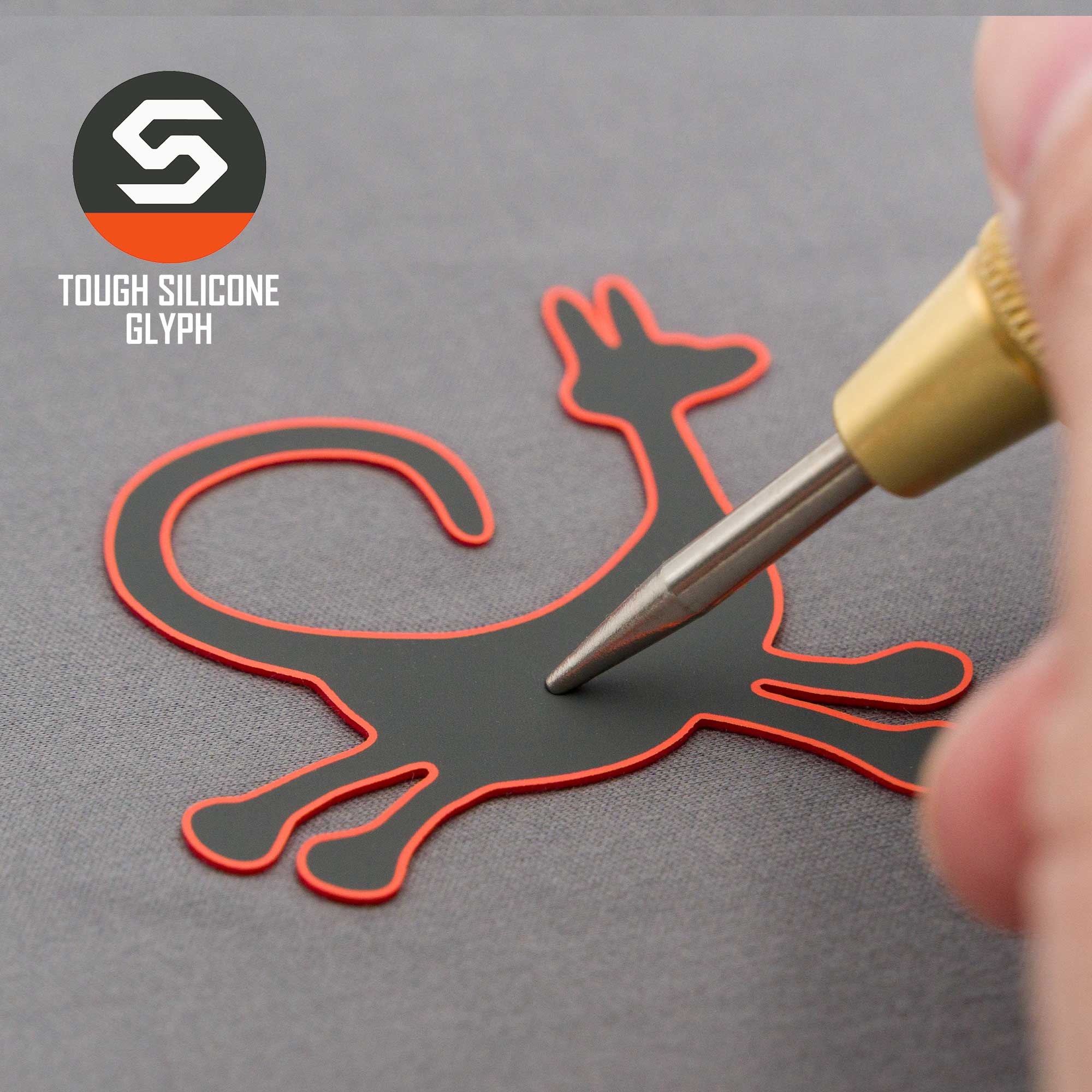 A hand presses a tool onto a flat, cat-shaped silicone patch with a red outline on a gray surface. The patch is the Eternal Glyph Moab Trickster Petroglyph (Silicone) for the Flexfit® Mesh Trucker Cap, inspired by Moab Utah designs.